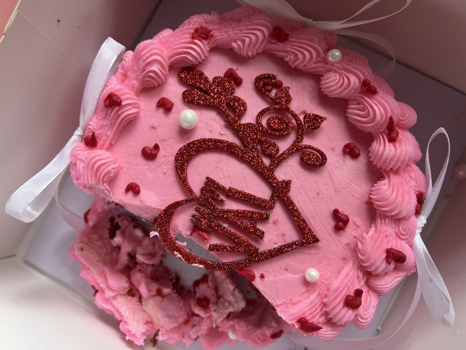 The image captures a top-down view of a light pink cake, partially eaten and presented within a white cake box. The cake is predominantly pink, covered in a smooth, light pink frosting on top, with decorative piped borders along its edges. These borders feature intricate, ruffled patterns, also in light pink frosting, interspersed with small, deep red heart-shaped sprinkles.
Two white, pearl-like spherical candies are visible on the cake's surface, strategically placed among the red sprinkles. The most prominent decoration is a large, glittery red cake topper positioned centrally on the cake. This topper features a heart shape at its base, within which appears to be a stylized depiction of two hands reaching towards or intertwined with each other. Extending upwards from the heart are ornate, vine-like branches with small leaf-like elements, all rendered in sparkling red glitter.
A significant portion of the cake, a large wedge or slice, has been removed from the lower-left section, revealing the cake's interior. The exposed crumbs are a mix of light pink and reddish hues, suggesting a red velvet or strawberry-flavored cake. The texture of the exposed cake is moist and crumbly.
The cake is nestled inside a pristine white cardboard box, which features white satin ribbons tied around its sides, suggesting it's either a gift or part of a special presentation. One ribbon is visible on the left side, tied in a neat bow, and another on the right side, also tied. The bright and even lighting illuminates the cake and its details clearly. The overall aesthetic is sweet and festive, likely indicating a celebration such as Valentine's Day or an anniversary.