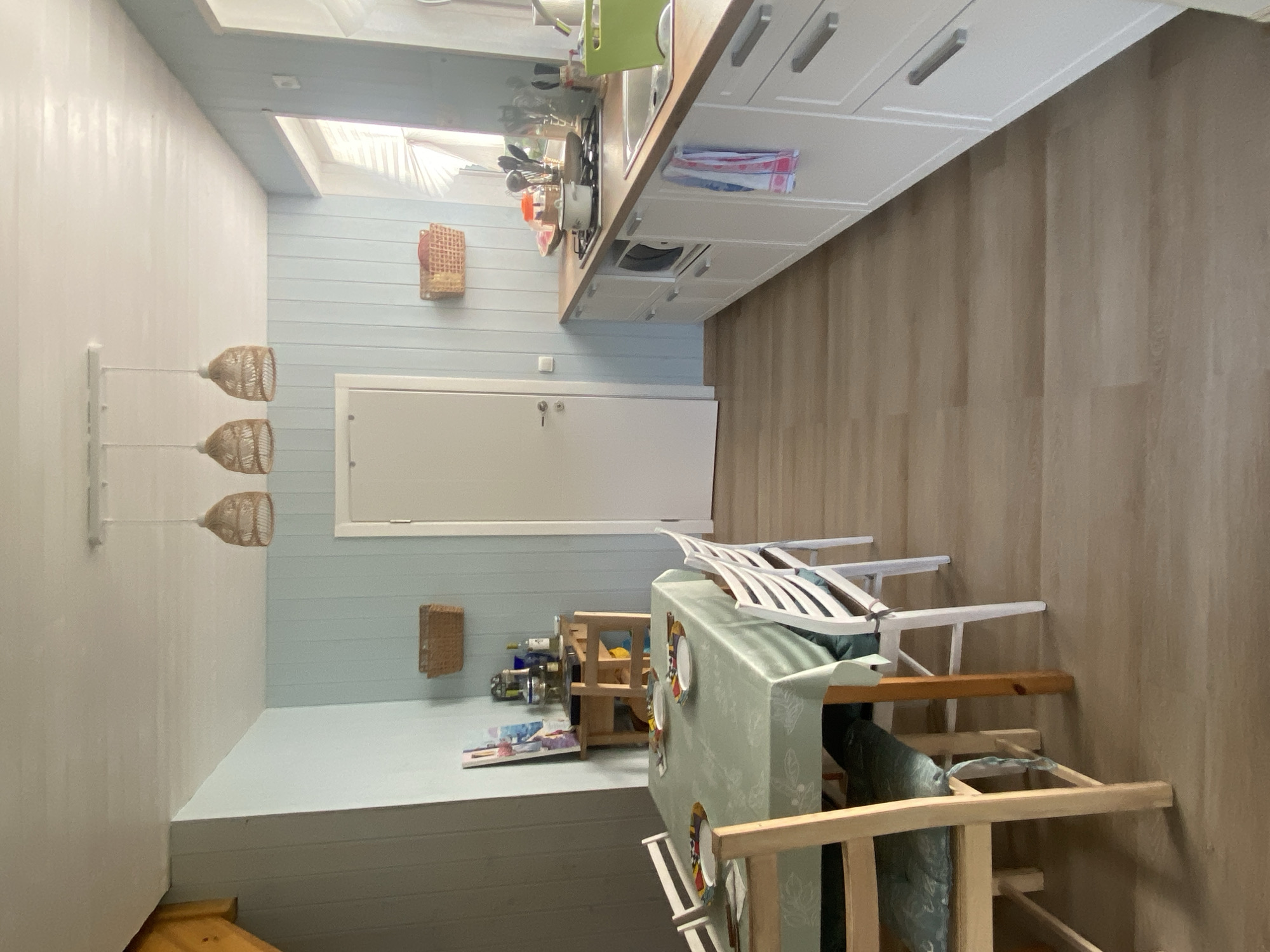 The image shows a bright and airy kitchen and dining area. The walls are paneled with light blue boards, and the floor is covered with light brown laminate flooring. A dining table covered with a light green tablecloth is set with plates and cutlery. Four chairs surround the table, two with light wood frames and cushions, and two with white frames. A small wooden shelf unit with various items is placed near the corner. In the background, a white door is visible. The kitchen counter is lined with white cabinets and a wooden countertop. A stove with a pot is on the counter, along with utensils and other kitchen items. A window with blinds is visible on the right side of the kitchen. Three woven pendant lights hang from the ceiling above the dining area. The scene appears to be a residential kitchen in Leipyasuo, Russia.