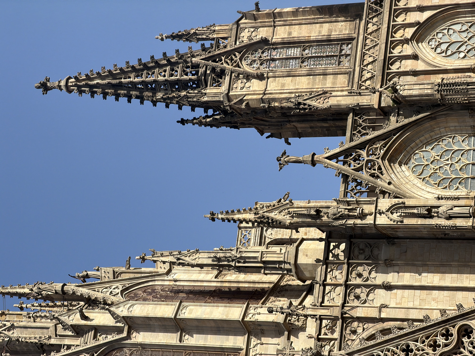 The image shows a close-up view of the Barcelona Cathedral in Barcelona, Spain, under a clear blue sky. The cathedral's Gothic architecture is detailed, with ornate stonework, spires, gargoyles, and stained-glass windows visible. The focus is on the upper portions of the building, emphasizing its height and intricate design. A small statue of an angel with wings is perched on one of the spires. The stonework is a warm, sandy color, illuminated by sunlight, creating shadows that highlight the architectural details. No people are clearly visible in the foreground, but the scale of the structure suggests it is a significant public space. The environment is clearly urban, with the cathedral being the dominant feature.