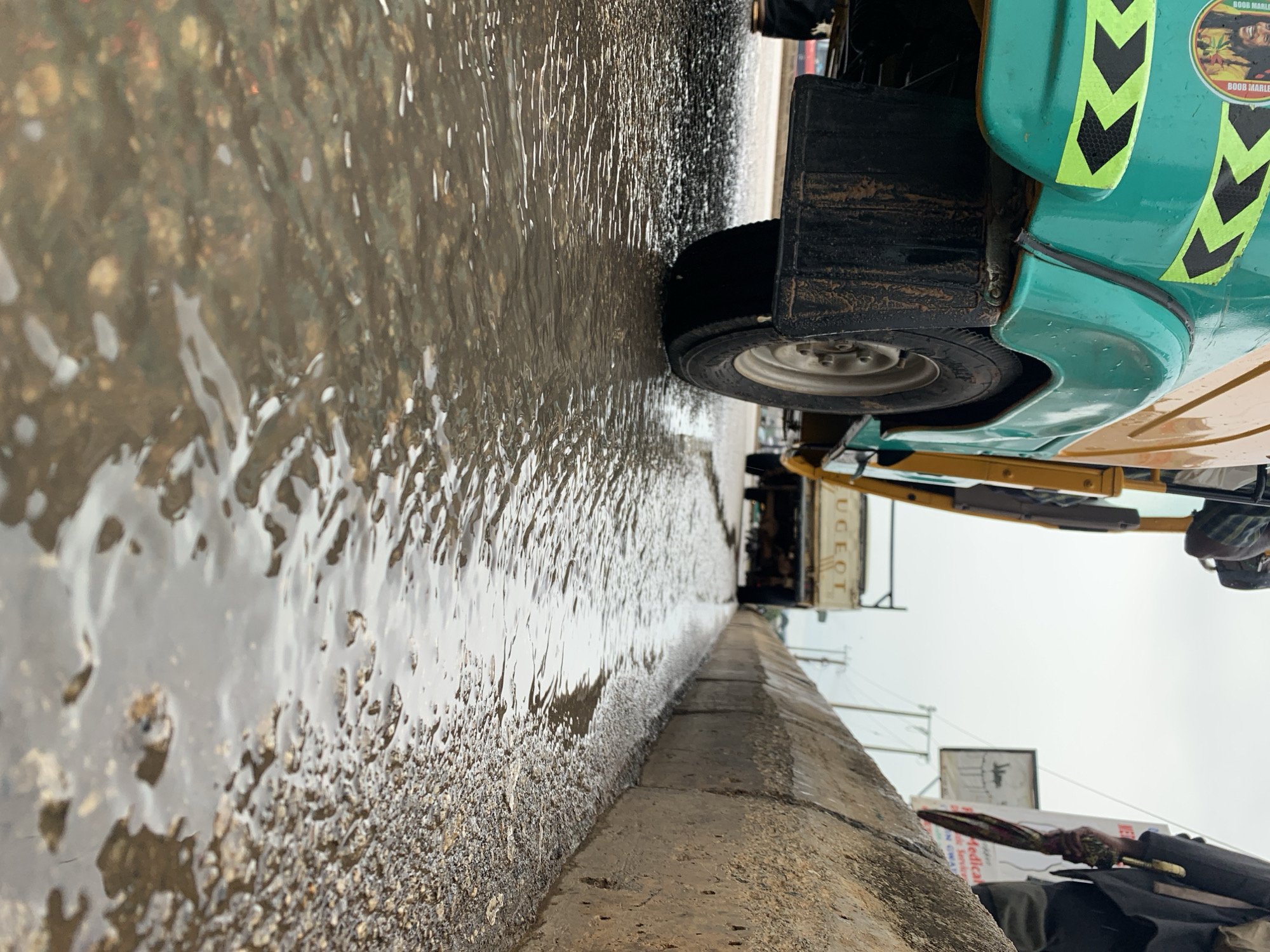 The image, oriented vertically, captures a street scene in Jos, Nigeria, featuring a large, reflective body of water dominating the foreground and midground, likely a flooded road after rainfall.
A green and yellow auto-rickshaw, commonly known as a keke napep in Nigeria, is the most prominent subject, appearing to be partially submerged in or driving through the extensive puddle on the left side of the frame. Its rear tire, mudguard, and a section of its body are clearly visible. The mudguard is dark and appears caked with mud. The vehicle's green body features black and yellow chevron warning stripes. A circular sticker near the rear wheel well displays a portrait of Bob Marley with the text "BOB MARLEY."
The water is murky, reflecting the overcast sky and the vehicle, and shows ripples suggesting movement. Small stones and debris are visible beneath the surface, especially closer to the camera. A concrete curb runs diagonally across the image, separating the flooded road from a higher area. On this higher ground, to the right, a person's arm and torso are partially visible, holding a long, slender object that could be an umbrella or a staff, suggesting navigation through the wet environment.
In the blurred background, other elements of a busy street are visible: a white vehicle with "PEUGEOT" partially discernable on its side, utility poles, and faint signs, one of which reads "Medical." The overcast sky indicates daytime conditions, and the widespread standing water points to recent or ongoing rain.