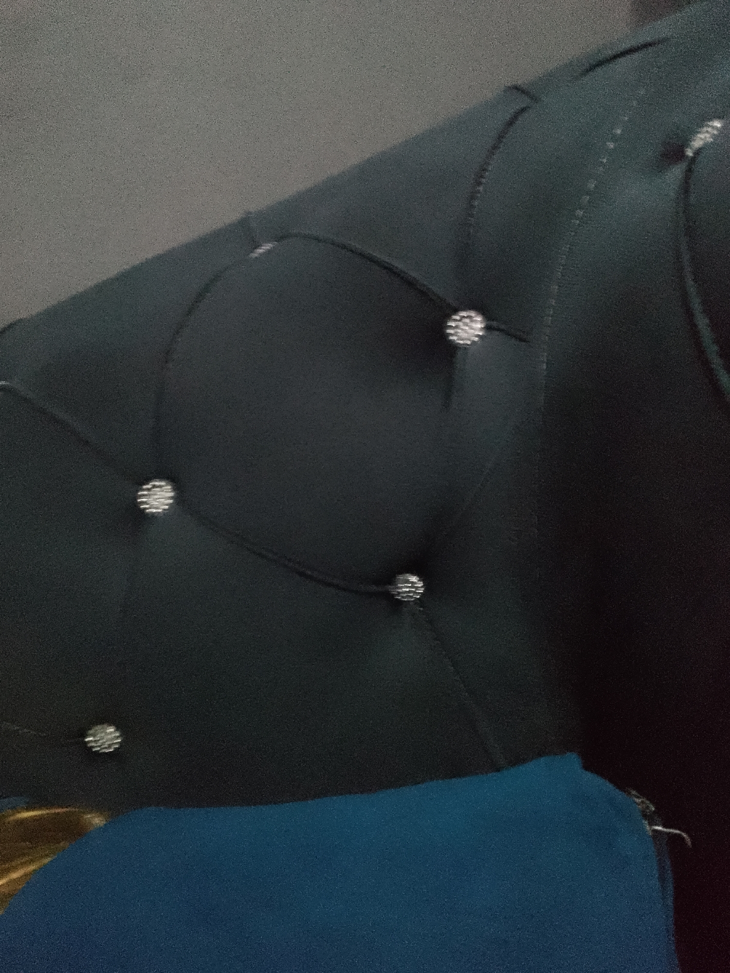 The image shows a close-up of a dark teal upholstered surface, likely a piece of furniture like a headboard or sofa. The upholstery features tufted detailing, with small, round, sparkling embellishments at the points where the fabric is pulled inward. These embellishments appear to be faceted, reflecting light. The fabric itself has a slight sheen and shows creasing and folds, indicating its texture and how it conforms to the underlying structure. In the foreground, a section of bright blue fabric is visible, appearing to be a separate item or part of the same furniture piece. The background is out of focus and appears to be a neutral gray, possibly a wall. The lighting is dim, creating shadows that highlight the depth of the tufting. The scene is static and does not depict any people or actions. There is no discernible information within the image that would allow for the identification of its location in Jauro Yinu, Nigeria, or any other specific geographical place.