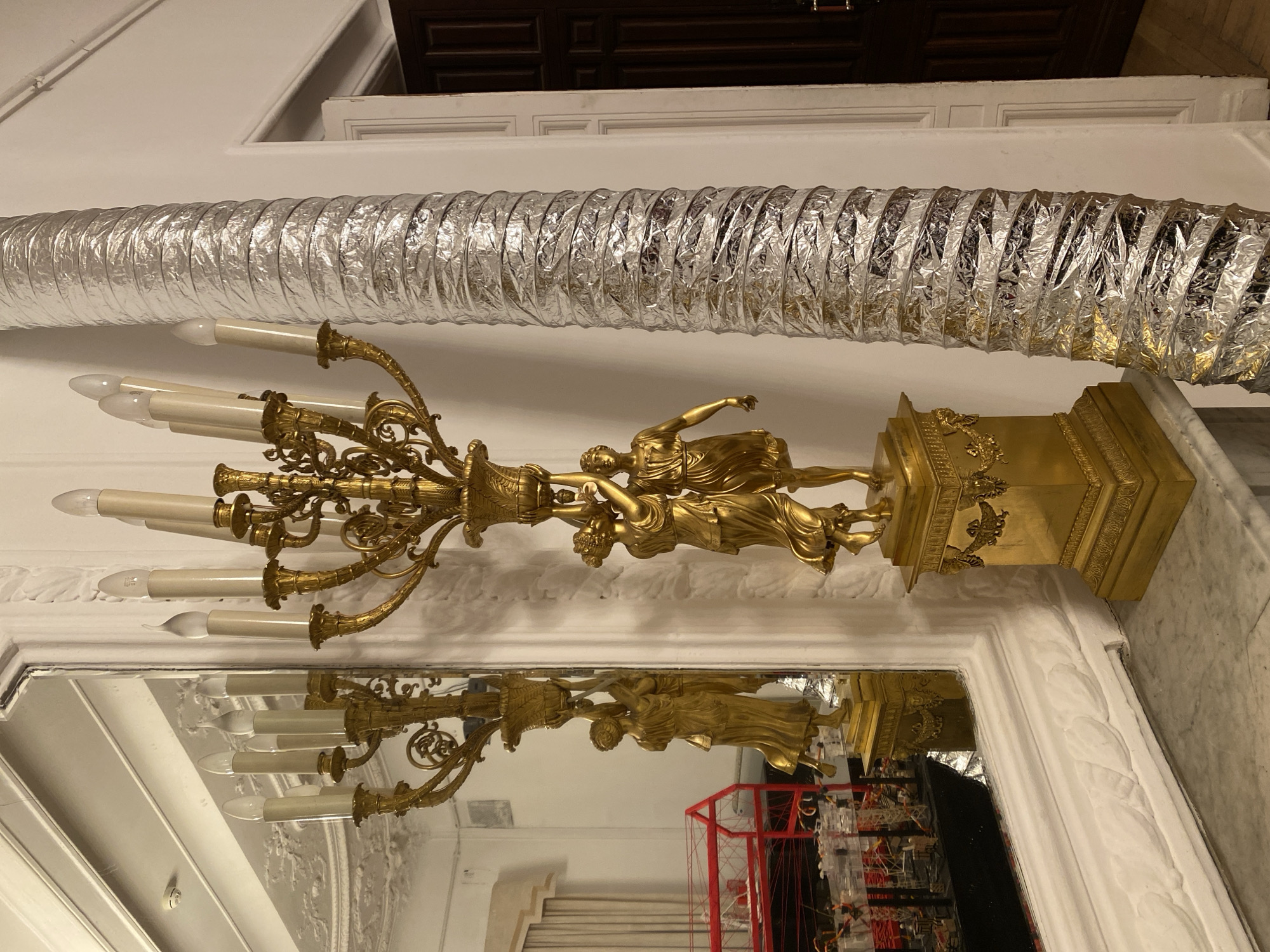 A gilded candelabra with sculpted figures sits atop a mantle in front of a mirror. A silver corrugated duct runs across the ceiling above the fixture. The mirror reflects the candelabra, interior of the room and stairs. The room is located in Moscow, Russia.