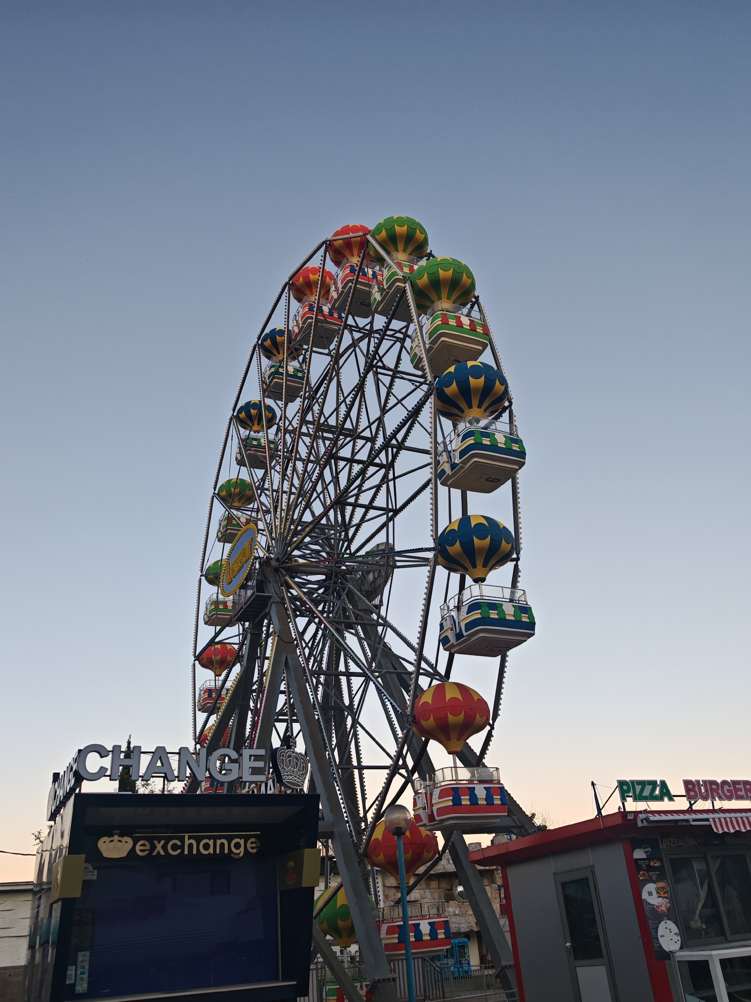 A Ferris wheel is centered in the shot, its gondolas designed to resemble hot air balloons in various color schemes: red and yellow, green and yellow, and blue and yellow. Below the Ferris wheel are commercial establishments. A "CHANGE" booth is on the left, with a sign underneath that reads "exchange" in a smaller font. To the right is a "PIZZA BURGER" stand. The sky above is a clear, light blue. The location is likely the city of Nesebar in Bulgaria, known for its Ferris wheel and other tourist attractions. There are no people visible in the image. The overall environment suggests a commercial area in a tourist destination.