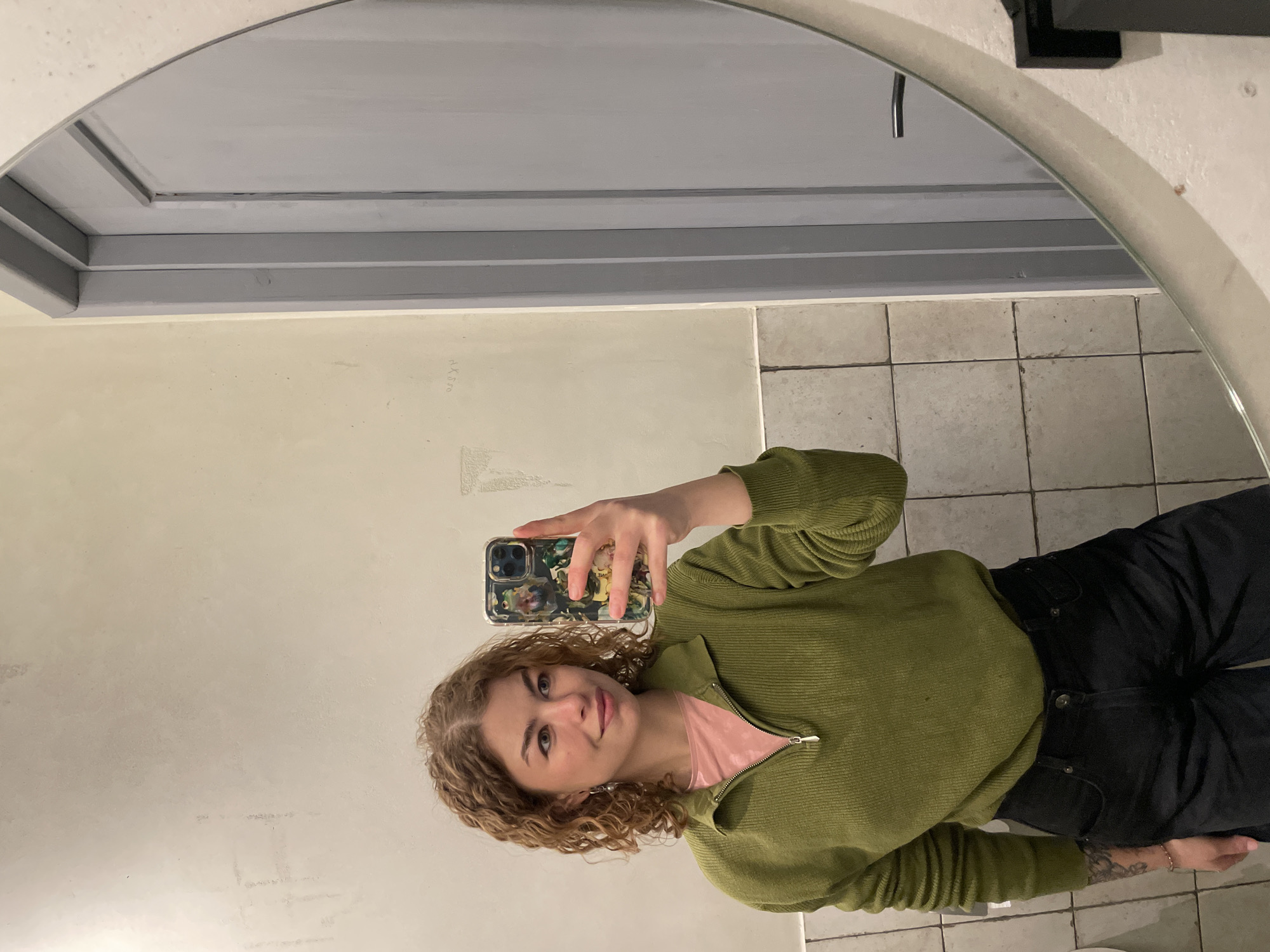 A woman is taking a selfie in a bathroom mirror. The mirror is oval-shaped. The wall is a light beige or off-white. There is a row of gray trim near the top of the wall. The floor is tiled. The woman has curly hair and is wearing a green sweater and black pants. She is holding a cell phone with a patterned case. I cannot infer the location based on the visual information provided.