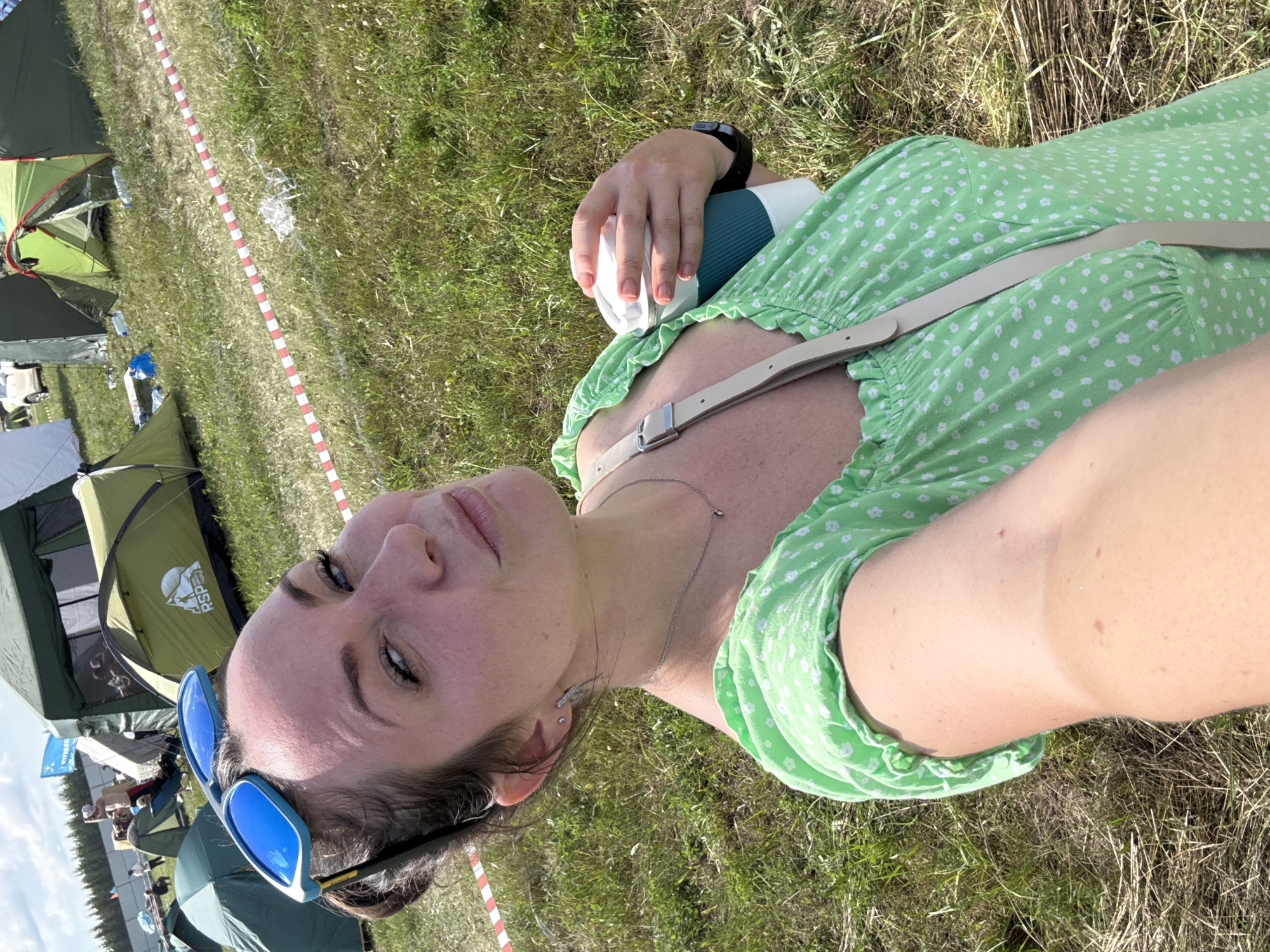 The image shows a woman taking a selfie outdoors, holding a disposable coffee cup in her right hand. She is wearing a green floral-print dress and blue sunglasses perched on her head. Behind her, several tents are visible, suggesting a campsite or festival setting. The ground is covered in dry grass and some red and white striped tape is present. The sky is bright and appears to be daytime. The woman is looking towards the camera with a slight smile. The scene is likely a recreational area given the tents and open space.