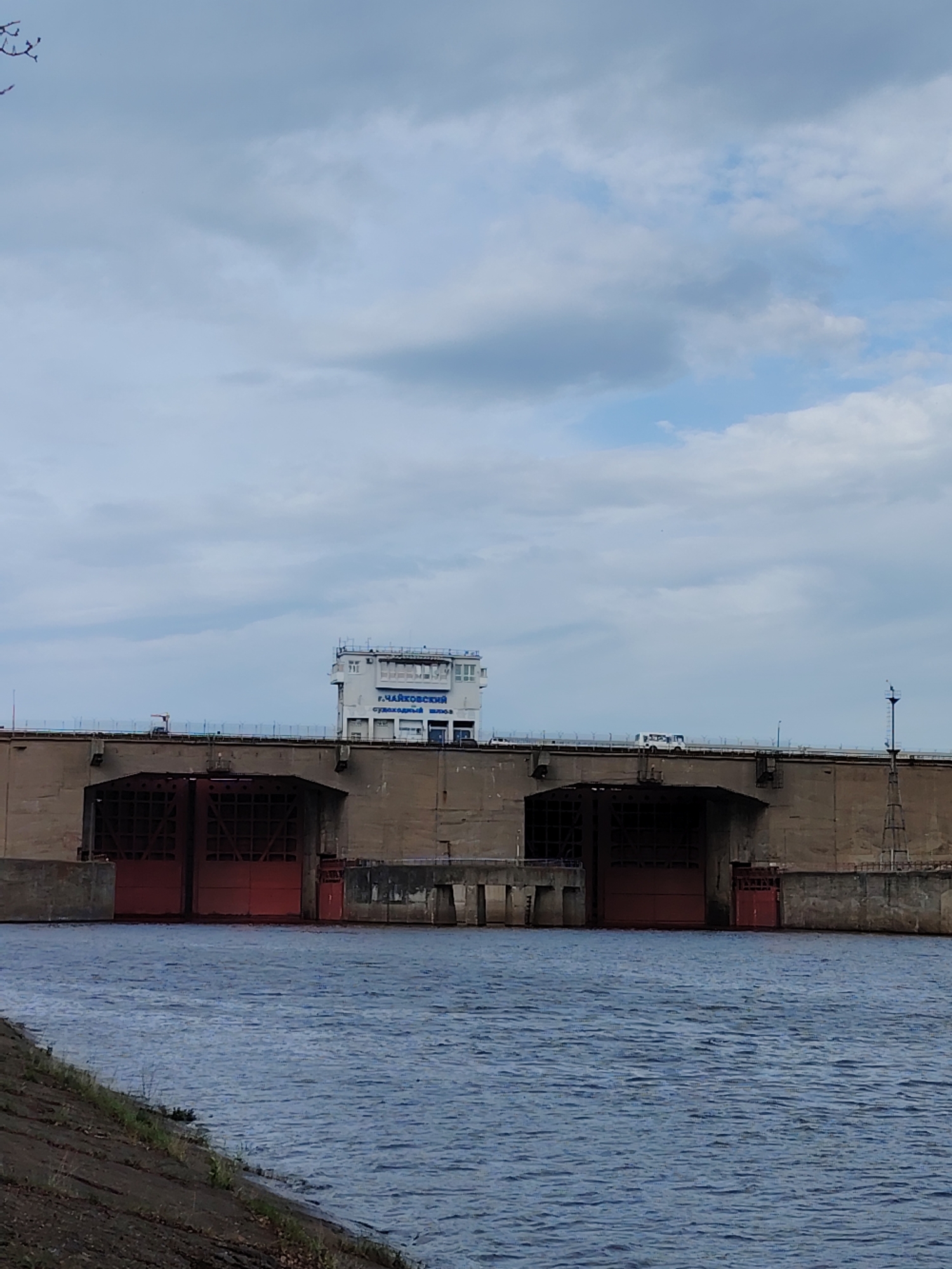 The image shows the Chaikovsky Dam in Chaikovsky, Russia. The dam has two large openings with red gates. A control tower sits atop the dam structure. A white bus is traveling across the dam. The foreground shows the water with some vegetation and a concrete embankment. The sky is cloudy.