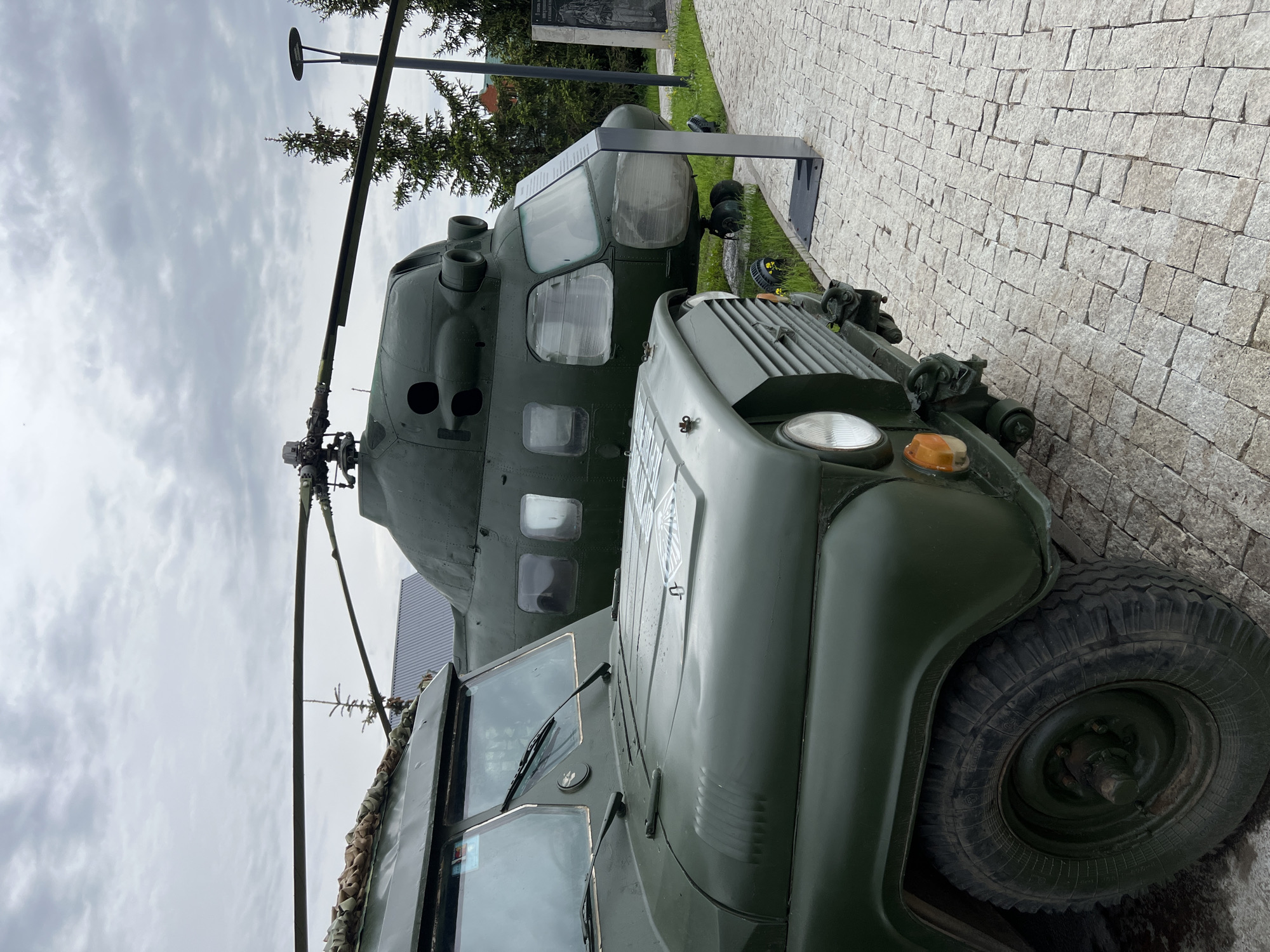 The image shows a Soviet Mil Mi-24 helicopter and a UAZ-469 military vehicle, both painted in olive drab, exhibited outdoors in the city of Введенская Слобода, Russia. The helicopter features its characteristic tandem cockpit, twin exhaust nozzles, and a prominent rotor blade. The UAZ-469, with its utilitarian design, includes a spare tire mounted on the hood and a canvas covering. The vehicles are displayed on a paved area with a cloudy sky in the background.