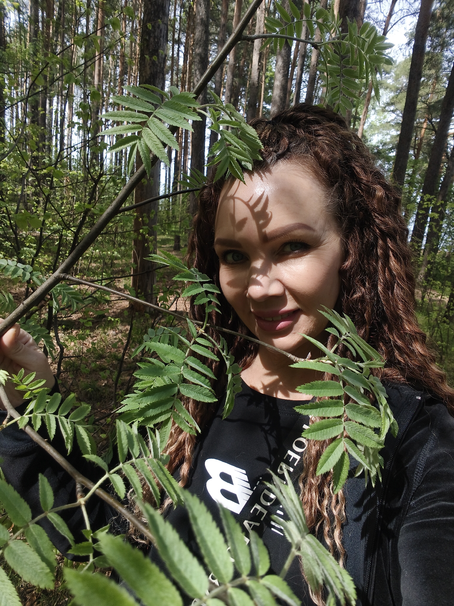 A woman with curly brown hair is standing in a forest. She is holding a branch with green leaves. The background consists of various trees and vegetation, with sunlight filtering through the branches.