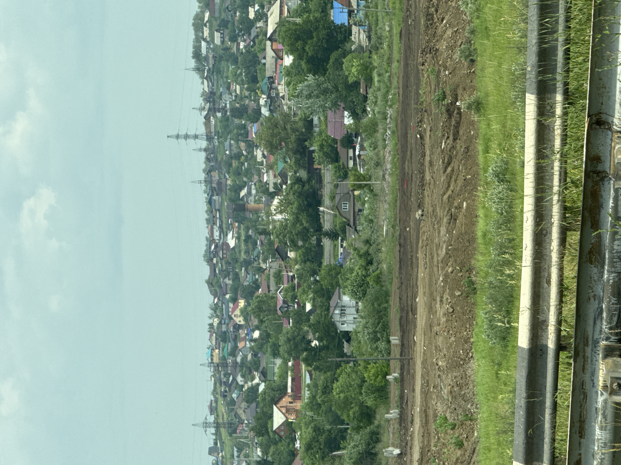 The image depicts a suburban hillside community in Solontsy, Russia, with a clear sky and scattered clouds overhead. A dense collection of houses, varying in size and architectural style, covers the undulating terrain. Many of the houses are single-family dwellings with pitched roofs, some painted in vibrant colors like red and blue, while others are more muted. Green trees and foliage are interspersed throughout the residential area, suggesting a well-established, vegetated neighborhood. Prominent in the middle ground are several tall, lattice-like electricity pylons, indicating the presence of power infrastructure. In the foreground, a section of a highway barrier, painted with alternating black and white bands, separates the viewer from a grassy embankment and a cleared, dirt construction site with visible tire tracks and piles of soil. The overall scene suggests a developing or expanding residential area on the outskirts of a city. There are no people visible in the image.