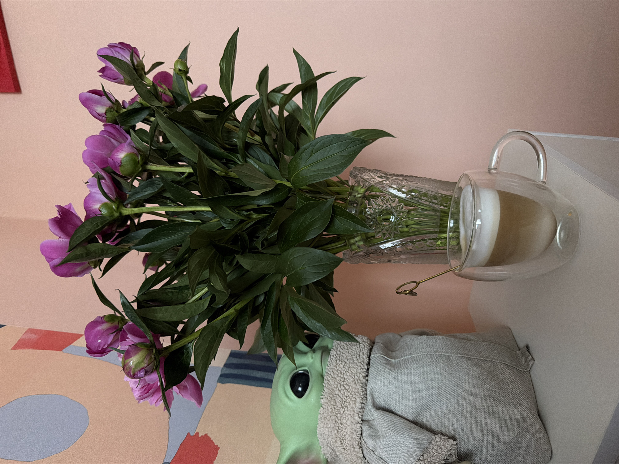 A still life scene depicts a vase of purple peonies arranged next to a double-walled glass mug containing a light-colored beverage. The vase is clear with an intricate cut glass design. The flowers are primarily purple, with some buds still closed. Abundant dark green leaves surround the flowers. The vase sits atop a green ceramic object, possibly a planter, partially covered with a light gray textured cloth. The mug is positioned on a white surface. The background consists of a light pink wall and a colorful patterned surface. The location is not discernible from the image.