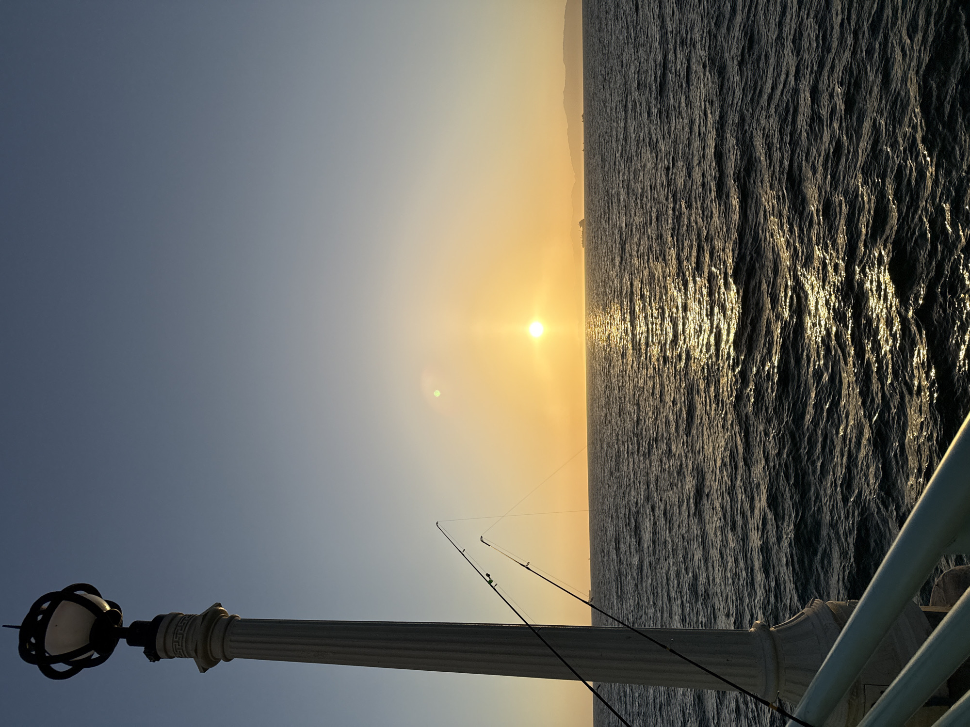 A vertical shot captures a sunset over the ocean from a pier in Manhattan Beach, California. In the foreground, a decorative lamppost stands to the left, with two fishing rods leaning against it. The rods are casting lines out over the water. The ocean occupies the majority of the frame, with gentle waves reflecting the warm glow of the setting sun. The sun itself is a bright orb near the center of the horizon, casting a golden path across the water. In the distance, a hazy shoreline with mountains is visible, along with a few silhouetted ships. The sky transitions from a deep blue at the top to a warm orange near the horizon, indicating the golden hour.