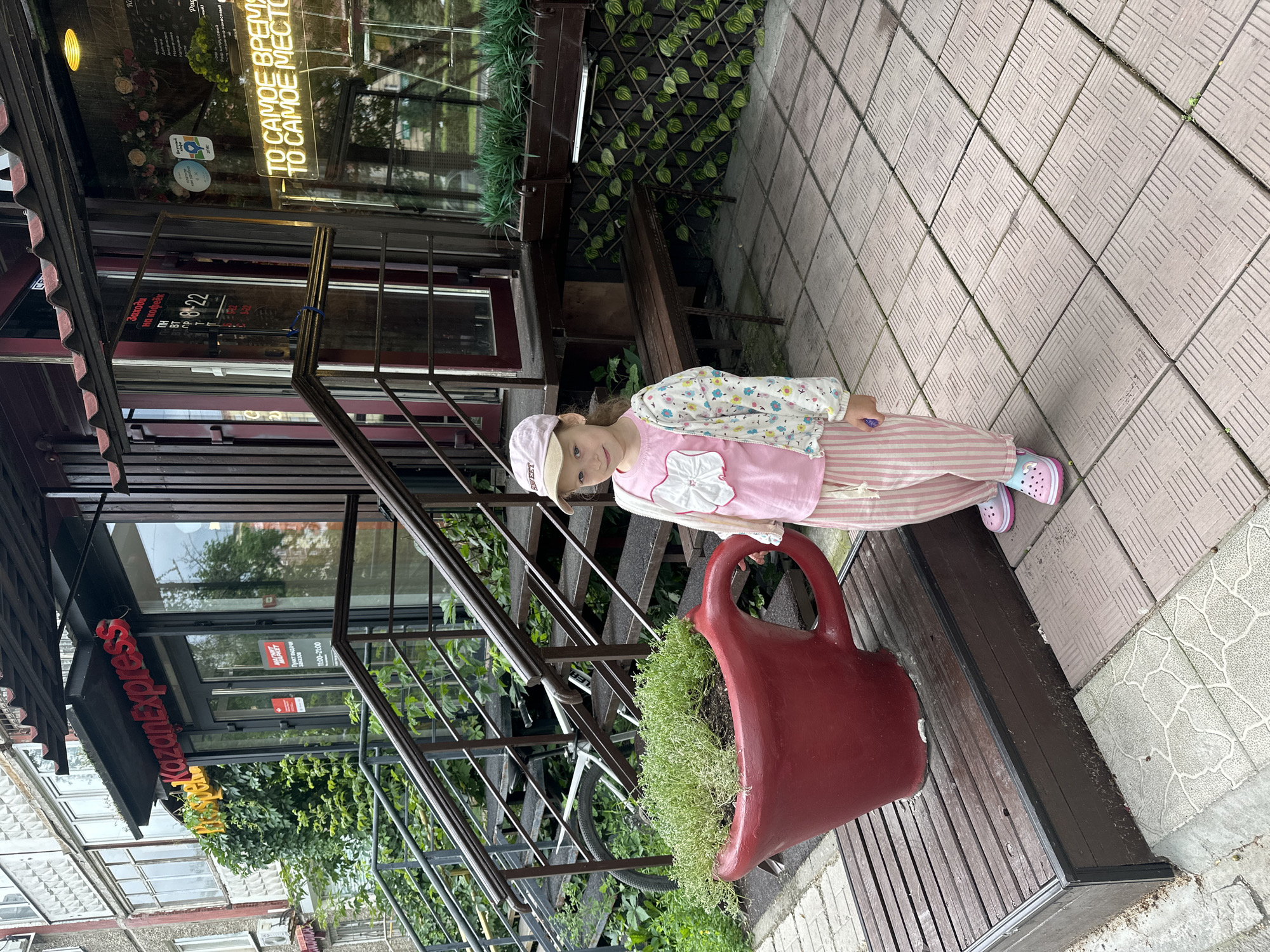 A young girl wearing a pink baseball cap, a pink t-shirt with a white bow, striped pink and white pants, and pink and blue crocs stands on a tiled patio in front of a building in Perm, Russia. She is looking at the camera. To her left, a large red cup-shaped planter overflows with green plants. Behind her, a staircase leads up to the entrance of a building with a sign that reads "KazanExpress." The building has large glass windows and a red awning. The area appears to be an outdoor seating area for a cafe or restaurant, with a wooden bench visible to the right of the girl. The ground is paved with square tiles.