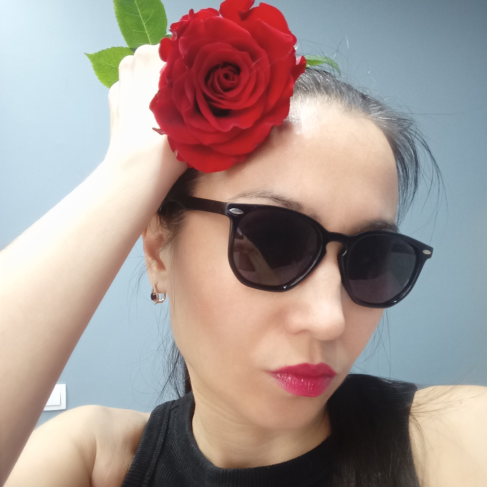 The image is a close-up portrait of a woman, taken from approximately the chest up, against a plain light blue or teal wall. The woman is looking down and slightly to her left, with her head tilted. Her left hand is raised, holding a vibrant red rose with green leaves positioned near her temple.
She has dark hair, which appears to be pulled back, with some loose strands around her hairline, including visible grey hairs. She is wearing black, Wayfarer-style sunglasses that cover her eyes. Her lips are made up with a bright, glossy, fuchsia-red lipstick. In her visible right ear, she wears a small, gold-toned hoop earring featuring a dark red gem. She is dressed in a black ribbed sleeveless top.
The setting appears to be indoors, with the light blue wall serving as a simple background. A white light switch is partially visible on the wall behind her left shoulder. The image has a well-lit, possibly daylight, indoor feel. The overall mood suggests a confident and stylish pose. There is no visible text in the image. The location context provided is Pervomaysky, Russia.