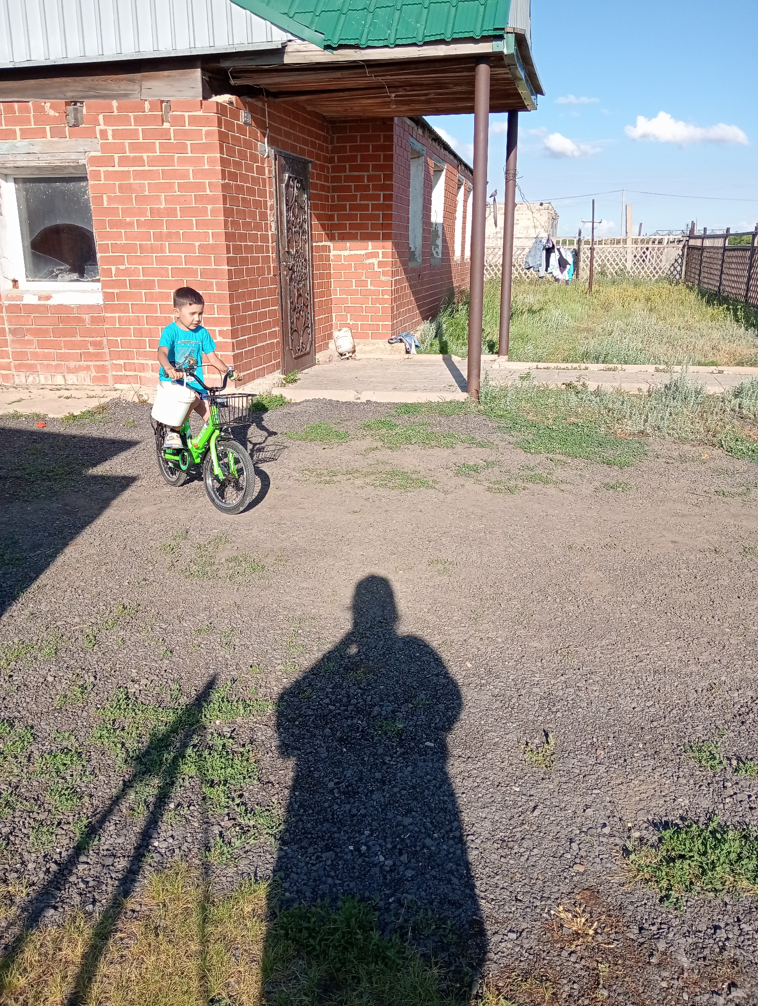A young boy in a blue t-shirt and shorts rides a bright green bicycle with a white basket and a black wire basket on the handlebars. He is on a gravel path in front of a brick building with a green metal roof. A shadow of the boy and his bicycle, as well as a larger shadow that appears to be an adult's, are cast on the path. The building has a window with broken glass and a dark, ornate metal door. To the right of the building, there is a grassy area with laundry hanging on a clothesline and a wooden fence. The sky is blue with scattered white clouds. The location appears to be a rural or semi-rural area in Kazakhstan.