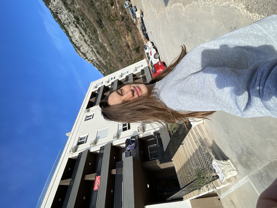 The image presents a scene in Budva, Montenegro, featuring a female individual taking a selfie outdoors under a clear blue sky. The individual has long brown hair and is wearing a grey crew-neck sweatshirt, with a slight smile on her face as she holds the camera.
Behind her, a multi-story building with a white facade and dark grey balconies is visible. A red sign, displaying "ИЗДАВANЬЕ" (FOR RENT) in Cyrillic, is affixed to a balcony on the second visible floor. Laundry, including a purple or blue item, hangs on a railing of a lower balcony. Several air conditioning units are mounted on the building's exterior. A set of light-colored stairs with dark railings leads up to the building's entrance, with a white decorative planter containing a small green plant positioned near the base of the stairs.
To the right of the building, a gravel and dirt parking area is observed, containing a red hatchback, a white SUV, a grey sedan, and a white utility truck. A small, light-colored dog is standing on the gravel near the parked vehicles. The background is dominated by a large, rocky mountain with sparse vegetation, ascending under the bright sunlight which casts distinct shadows across the scene.
