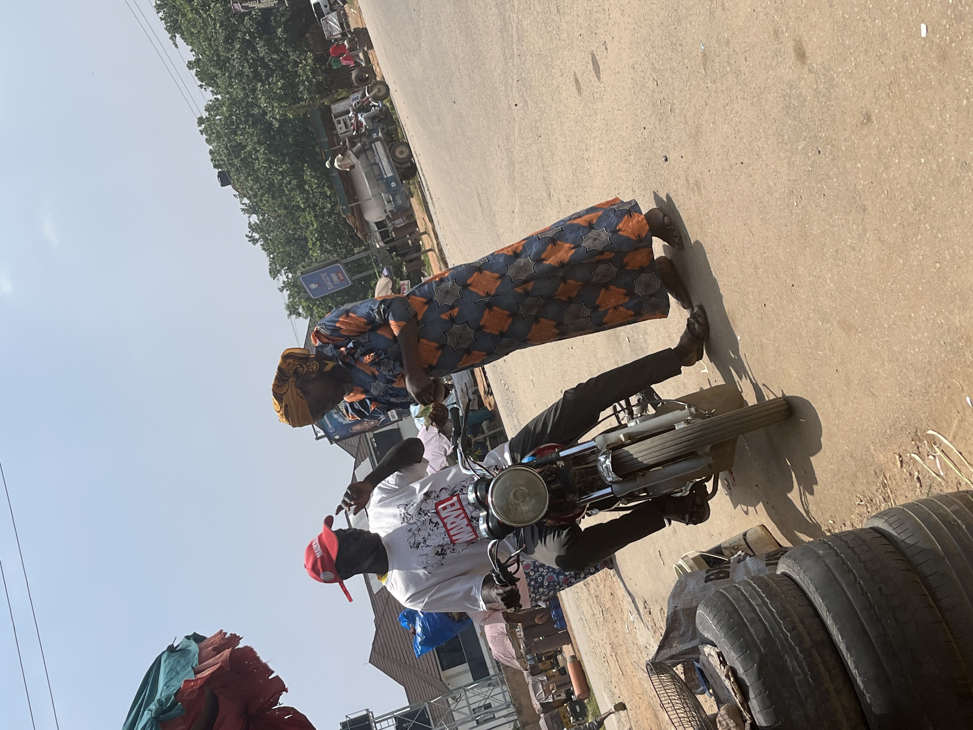 A man wearing a white t-shirt with a Marvel logo, black pants, and a red baseball cap is seated on a motorcycle. A woman in a long, patterned dress and headscarf stands next to him, her hand resting on the motorcycle. They appear to be in a bustling outdoor market or roadside area. In the background, there are other motorcycles, a truck, and various goods, suggesting a commercial setting. The scene is outdoors with clear sky, indicating daytime, likely sunny weather. The overall impression is one of everyday activity in a Nigerian urban or semi-urban environment. Some text is visible on the man's t-shirt, reading "MARVEL." A blue sign with white text is also partially visible in the background, but the text is illegible. The composition of the image is unusual, with the camera tilted, making the ground appear to be a vertical surface and the sky a horizontal one.