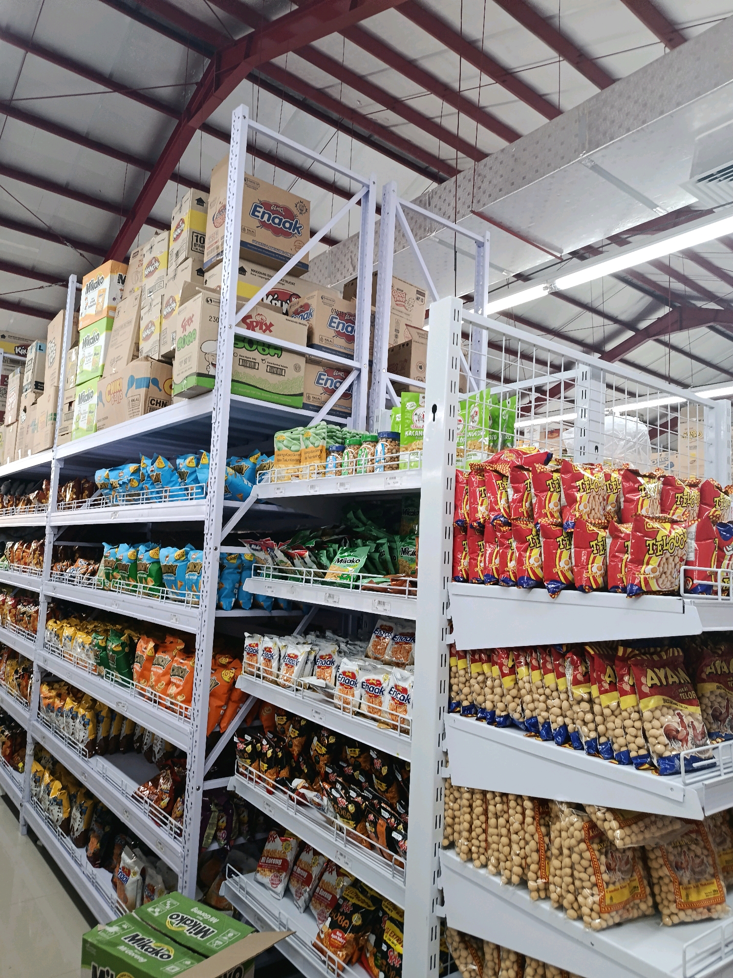 The image shows the interior of a supermarket in Bulukumba, Indonesia, with shelves stocked with various packaged snacks and food items. The shelving units are white metal, and the ceiling is a corrugated metal structure with exposed red beams and light fixtures. The aisle appears to be empty of people. The products on display include bags of what appear to be nuts or chips, many with colorful packaging featuring Indonesian text. Several boxes are stacked on the top shelves, some with branding such as "Enaak" and "Mikako". The overall impression is one of a well-stocked retail environment.