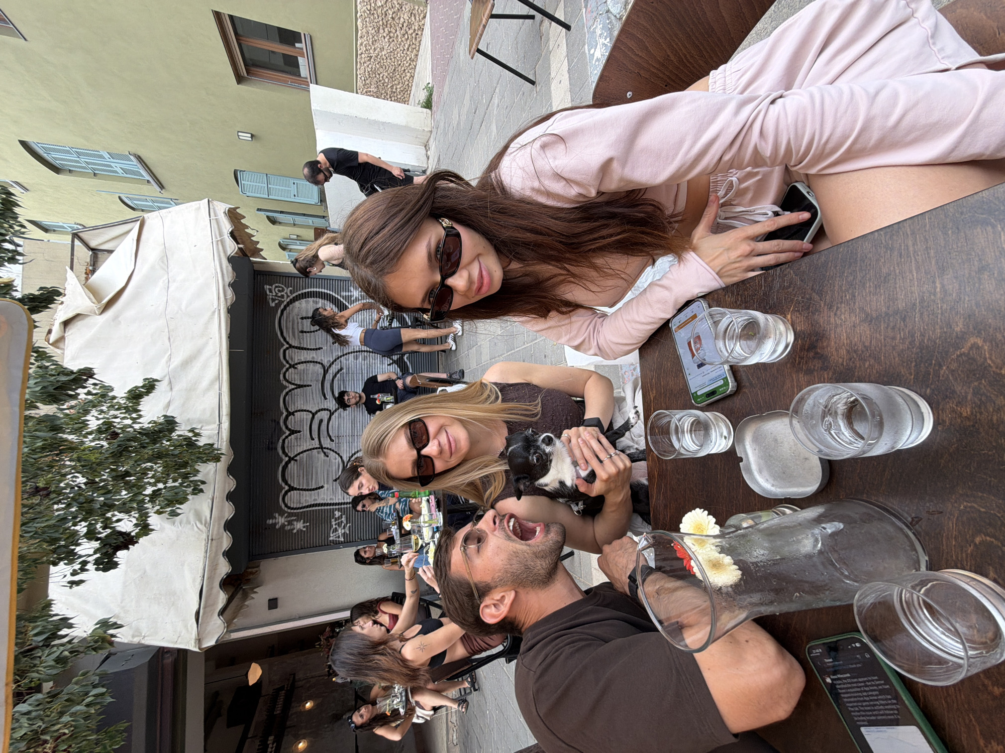 The image depicts a group of people gathered around a wooden table at an outdoor cafe in what appears to be an urban setting. The cafe is situated on a street lined with buildings, some featuring awnings and architectural details such as window shutters. Graffiti adorns a closed metal gate. The cafe is likely in Tel-Aviv, Israel. The group consists of approximately ten individuals, mostly young adults. One woman is wearing sunglasses and holding a small dog. One man is opening his mouth wide. There are several glass pitchers and water glasses on the table, along with mobile phones. The overall impression is a casual, social gathering.