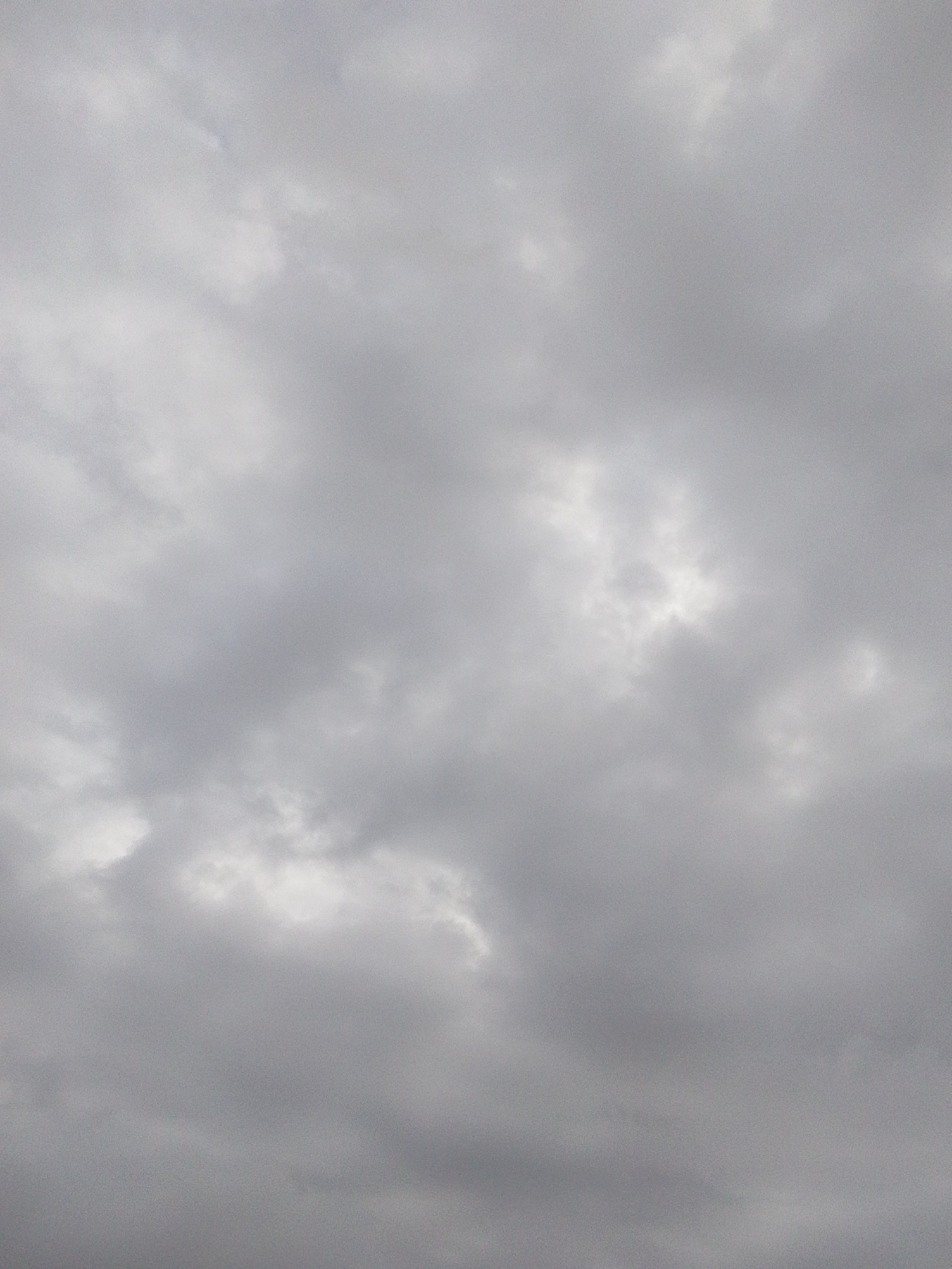 The image is a full-frame shot of a cloudy sky. The clouds are a uniform gray, with some areas appearing lighter due to sunlight filtering through. There is no discernible detail in the clouds, such as specific cloud formations or textures. The image is evenly lit, with no shadows or highlights that would indicate a specific time of day or weather condition beyond overcast. The scene is entirely natural, composed solely of the sky and clouds. No man-made objects, people, or any features of the city of Serti, Nigeria, are visible. The image is devoid of any action or interaction.
