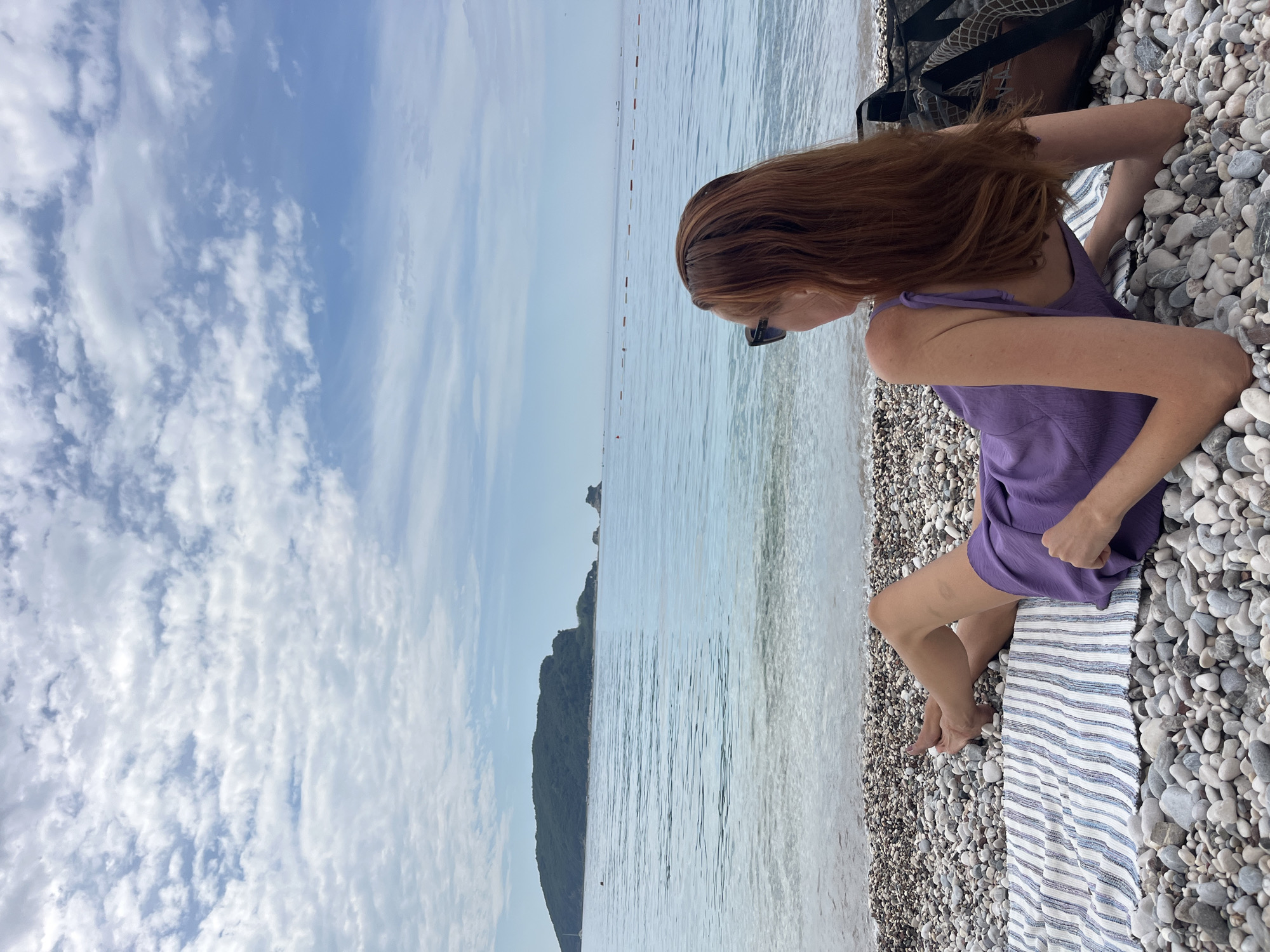 A woman with red hair, wearing a purple dress and sunglasses, is relaxing on a pebble beach in Budva, Montenegro. She is sitting on a blue and white striped towel. The beach consists of small pebbles. In the background, the sea is visible, with a small island on the horizon. The sky is light blue with scattered white clouds. A black tote bag rests behind her. Buoys are in the water and a mountain is along the coast.
