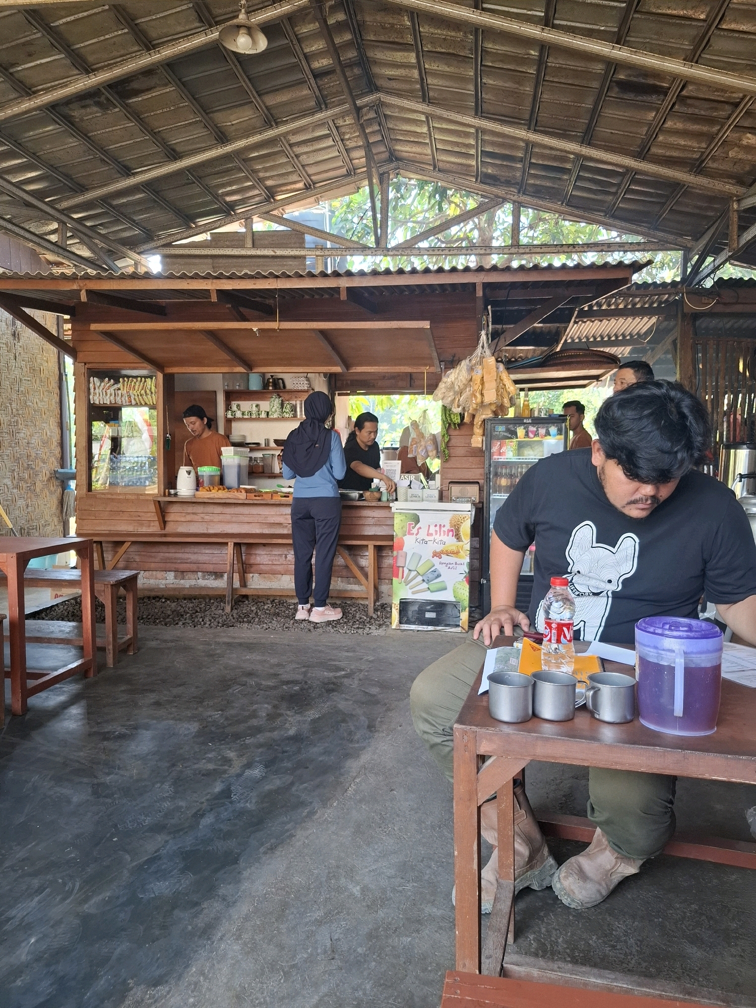 The image depicts an outdoor cafe or food stall in Cibentang, Indonesia, with a rustic, open-air setting under a corrugated metal roof supported by wooden beams. In the background, a wooden counter area is set up with shelves stocked with various food items and drinks. Three individuals are visible behind the counter, attending to customers or preparing orders.
In the foreground, a man wearing a black t-shirt with a graphic print sits at a wooden table, looking down at papers and a drink container. Two metal cups and a bottle of water are on the table in front of him. His posture suggests he is engaged in a task or examining something on the table. The cafe floor appears to be a dark, possibly concrete or asphalt surface, with some scattered gravel. The overall atmosphere is casual and functional, typical of local eateries in the region.