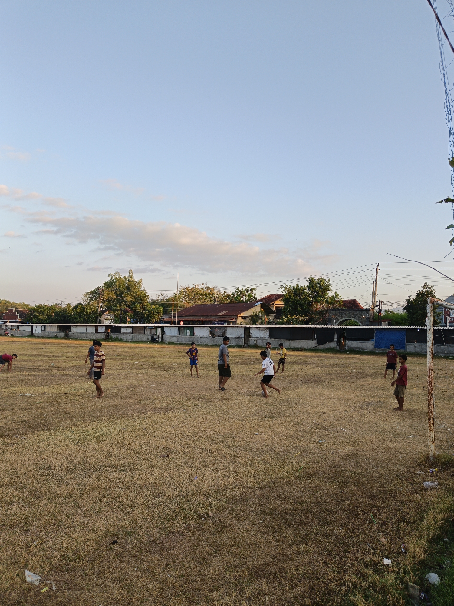 A group of young boys are playing soccer on a dry, grassy field in Trucuk, Indonesia. The sun is setting, casting a warm glow over the scene. In the background, there are trees, houses, and a volleyball net. The boys are dressed in casual clothing and appear to be enjoying themselves. The field is littered with some trash, including plastic bags and bottles.