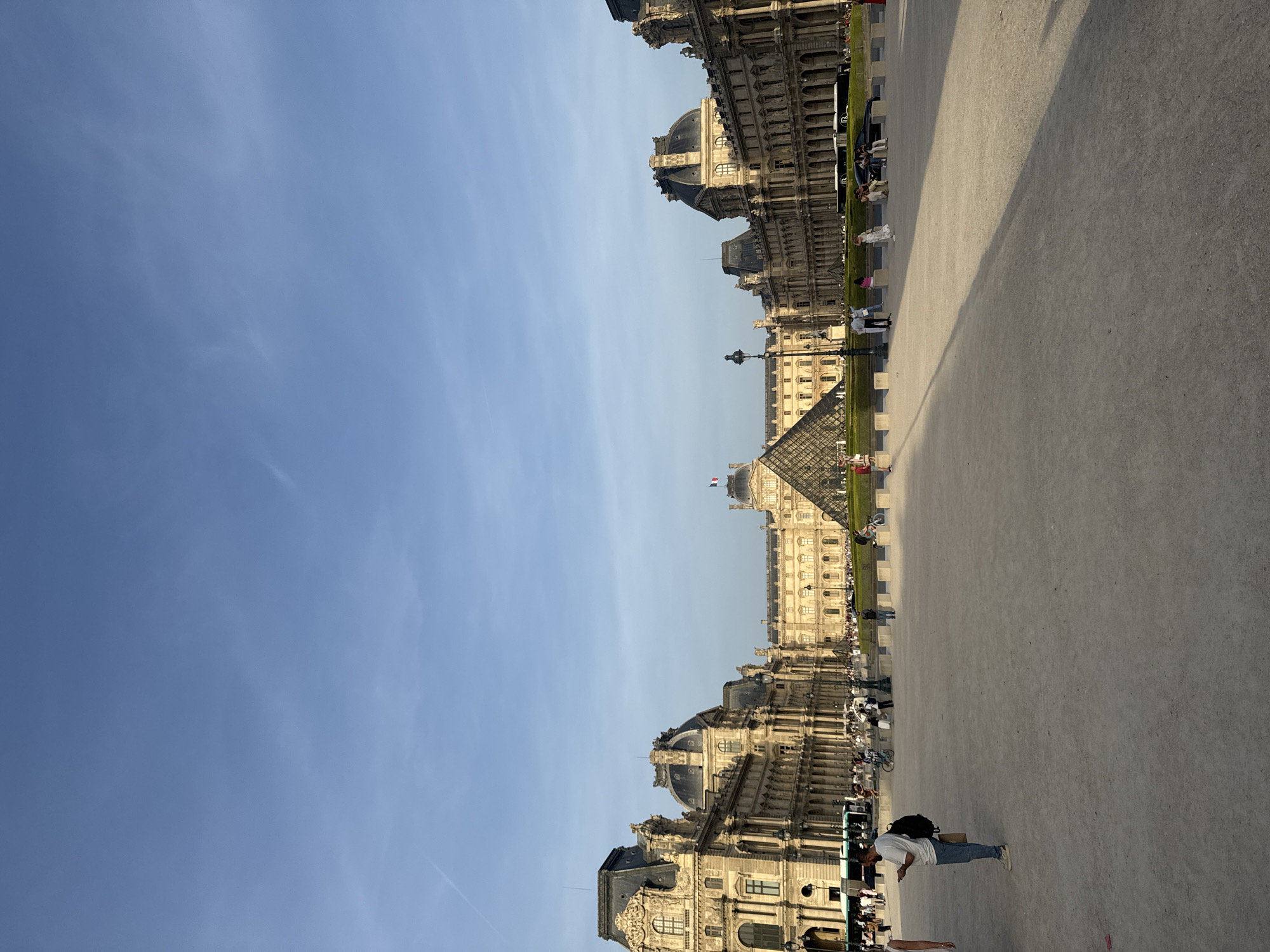 The image depicts the Louvre Museum in Paris, France. The iconic glass pyramid stands prominently in the courtyard, surrounded by the historic museum buildings. People are seen walking around the courtyard, and the scene is set against a clear blue sky. The overall atmosphere is typical of a sunny day at a popular tourist destination.