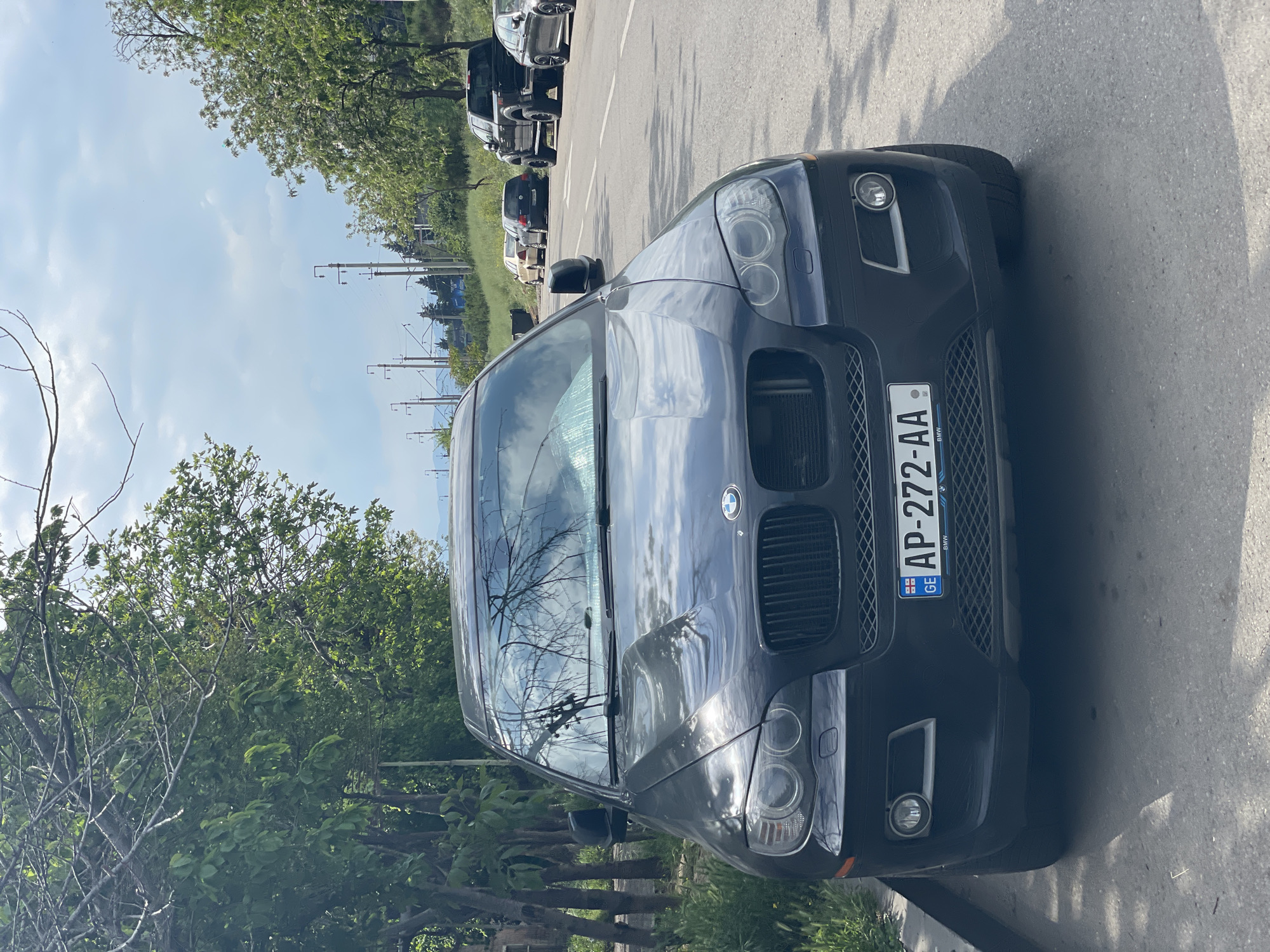 A parked BMW X6 is prominently displayed in the foreground, revealing its front grill, headlights, and the Georgian license plate AP-272-AA. A line of parked cars extends into the background along the road. The environment includes trees on the left and a sky visible behind the cars and through the windshield reflection. The image appears to be taken on a street in Tbilisi, Georgia.