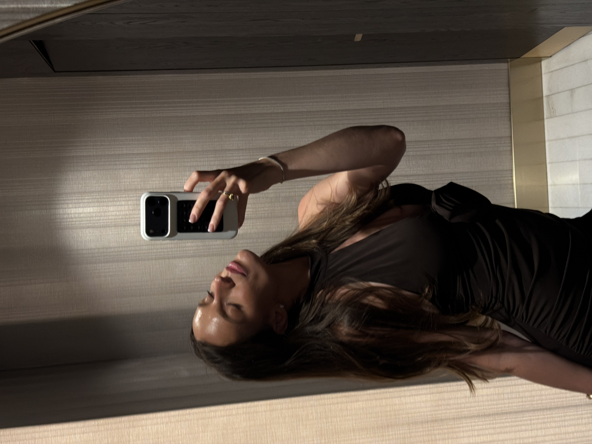 A female is depicted in a mirror selfie within an indoor setting in Budva, Montenegro. The subject, appearing to be in her 20s or 30s, possesses long, dark brown hair and a tanned complexion. She wears a black, sleeveless, high-neck dress featuring a frontal cutout and side ruching. Her head is tilted upward and to her left, eyes closed, with lips slightly parted, displaying light pink lip gloss. She holds a white smartphone with a dark camera module in her raised right hand. A gold-colored ring is visible on her right ring finger, and a slender, silver-toned bracelet adorns her right wrist. Her fingernails are manicured with white French tips. The background, a reflection in the mirror, consists of light beige, vertically textured wallpaper. To the right, a darker, vertically oriented panel or door is visible. A gold-colored baseboard runs horizontally at the bottom of the wall, above a white tiled floor. Lighting produces highlights on the subject's face and illuminates the textured wall, with some shadowing present on the right side of the frame.