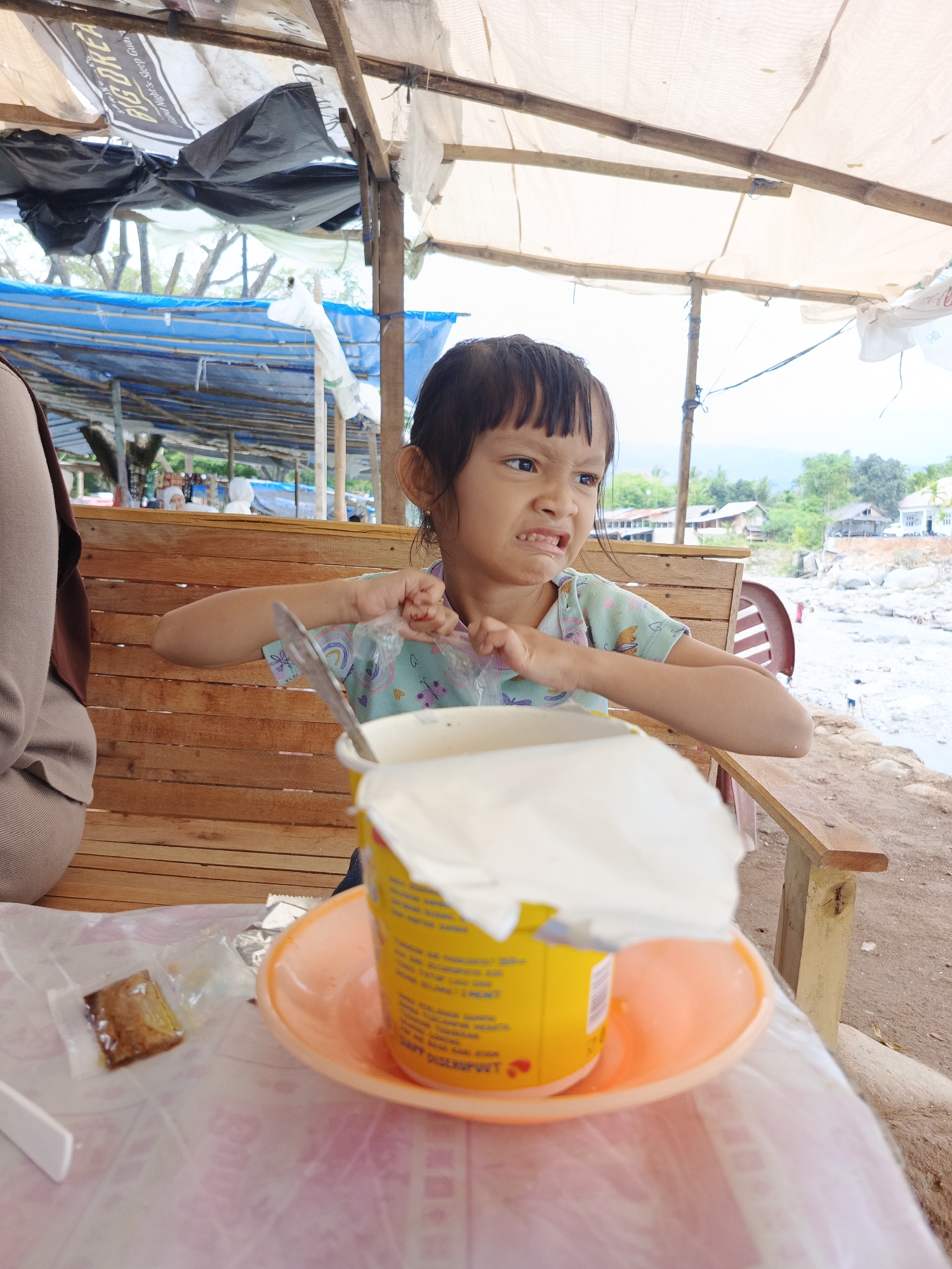 In Meurah, Indonesia, a young girl with dark bangs and hair, wearing a patterned light blue short-sleeved shirt, is seated on a light brown wooden bench at a table covered with a pink patterned plastic tablecloth. She exhibits a scrunched-up facial expression, indicative of concentration or mild displeasure, while manipulating a clear plastic packet with both hands above a yellow cylindrical container. The container, bearing visible text and a red heart icon, rests on an orange plastic saucer, with a white lid partially lifted or placed on top. Red nail polish is visible on the girl's fingernails. To her immediate left, the shoulder and upper arm of another person, clad in a beige garment, are partially visible. On the table, to the left of the container, a small, sealed transparent pouch containing a brown solid object is present, alongside a partially visible white utensil handle.
Overhead, an improvised canopy is constructed from multiple pieces of fabric, including white, blue, and black tarpaulins, supported by bamboo poles and rudimentary wooden beams. Text on a black banner within the canopy is partially discernible as "BIG GORE F". The background reveals an open-air setting, with the structures overlooking a wide, dry, rocky riverbed. Scattered buildings and dense green foliage are present on the opposing bank, with mountains in the distant horizon. Indistinct figures are visible near the riverbed in the background.
