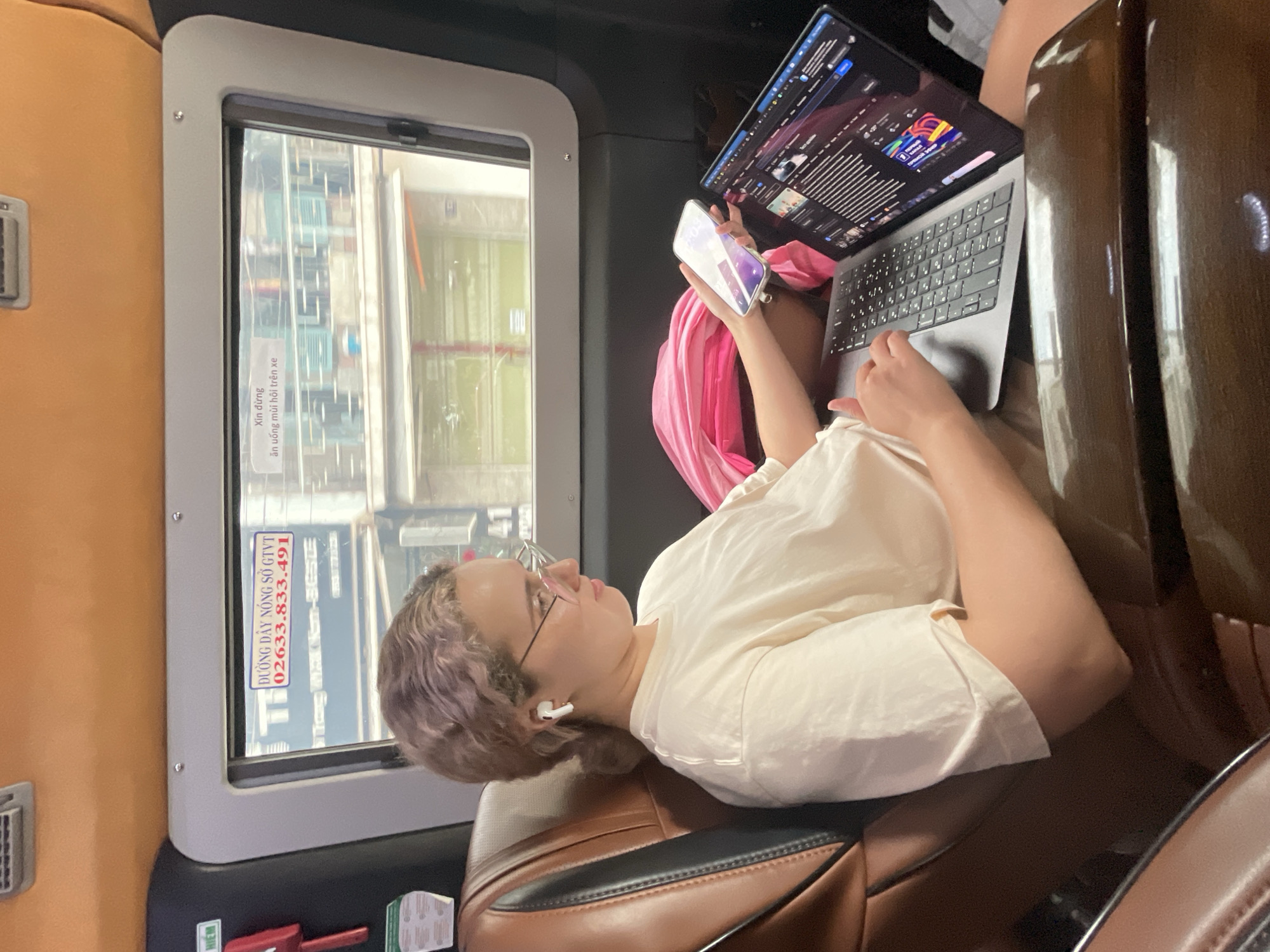 The image shows a woman seated in a vehicle, possibly a bus or private car. She has light purple hair and is wearing glasses and an earpiece. She's holding a smartphone in her left hand and using a laptop with her right hand. The laptop displays a screen with text and graphics. She is wearing a light-colored shirt and a pink scarf or garment is visible behind her.
Outside the vehicle's window, the scene suggests an urban environment, likely in Nha Trang Ward, Vietnam, as indicated by the Vietnamese text on a sticker on the window frame. The sticker displays a phone number and a reminder not to consume strong-smelling food or drinks on the vehicle.