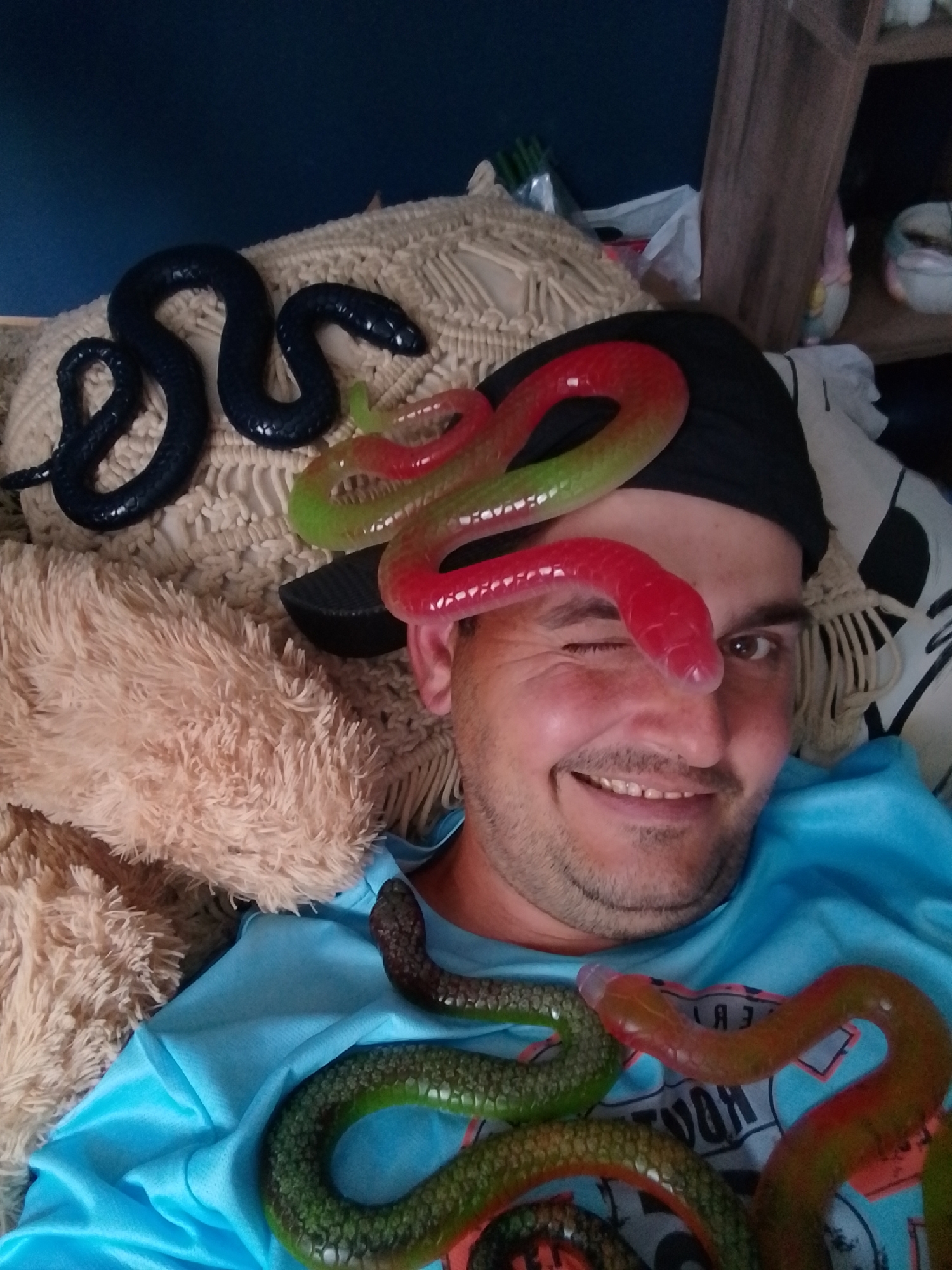 A man wearing a black beanie and a blue t-shirt is lying down, with several gummy snakes placed on and around him. One large black snake is coiled on a textured, light-colored cushion behind him. Two brightly colored gummy snakes, one red and green, the other red and orange, are draped across his face and body. Smaller, green and brown gummy snakes are on his chest. The background includes a dark blue wall, a shelf with various items, and a fluffy beige pillow. The man is smiling and winking, with one gummy snake positioned over his closed eye. The scene appears to be indoors, likely in a residential setting.