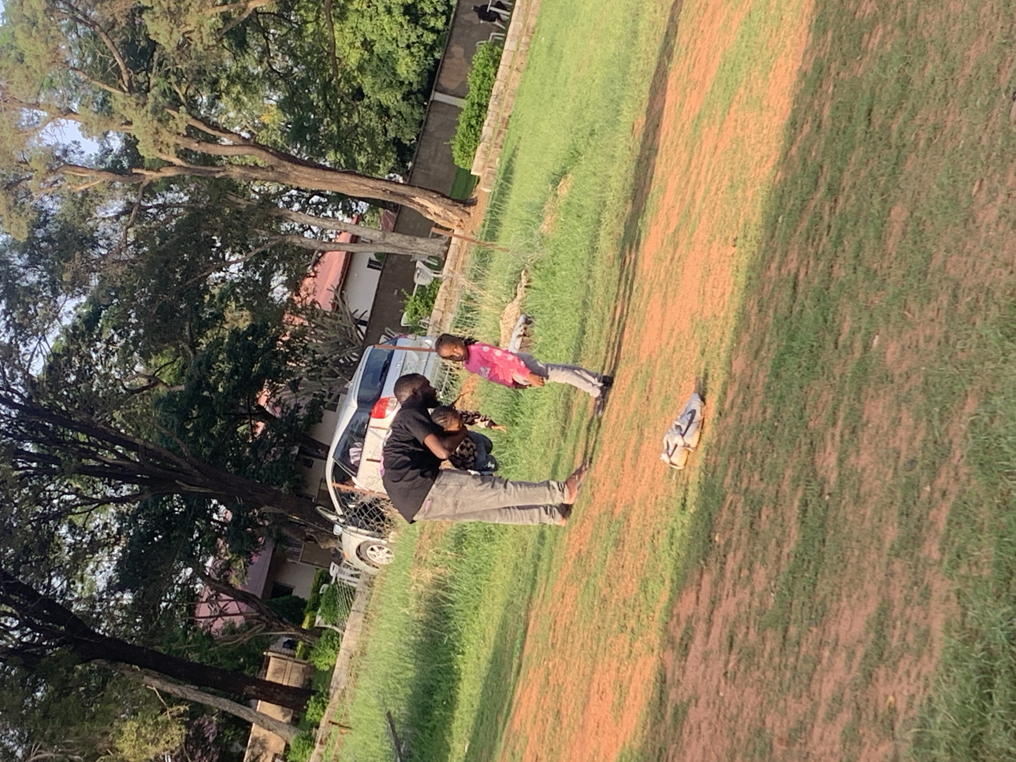 A Black man wearing a black t-shirt and gray pants is holding a young Black child wearing a pink and white polka-dot shirt and gray pants, who is looking at the man. They are standing on a dirt and grass area in front of a grassy embankment with trees and a building in the background. A pair of white sneakers are on the ground in front of them. The environment appears to be an outdoor space in Jos, Nigeria.