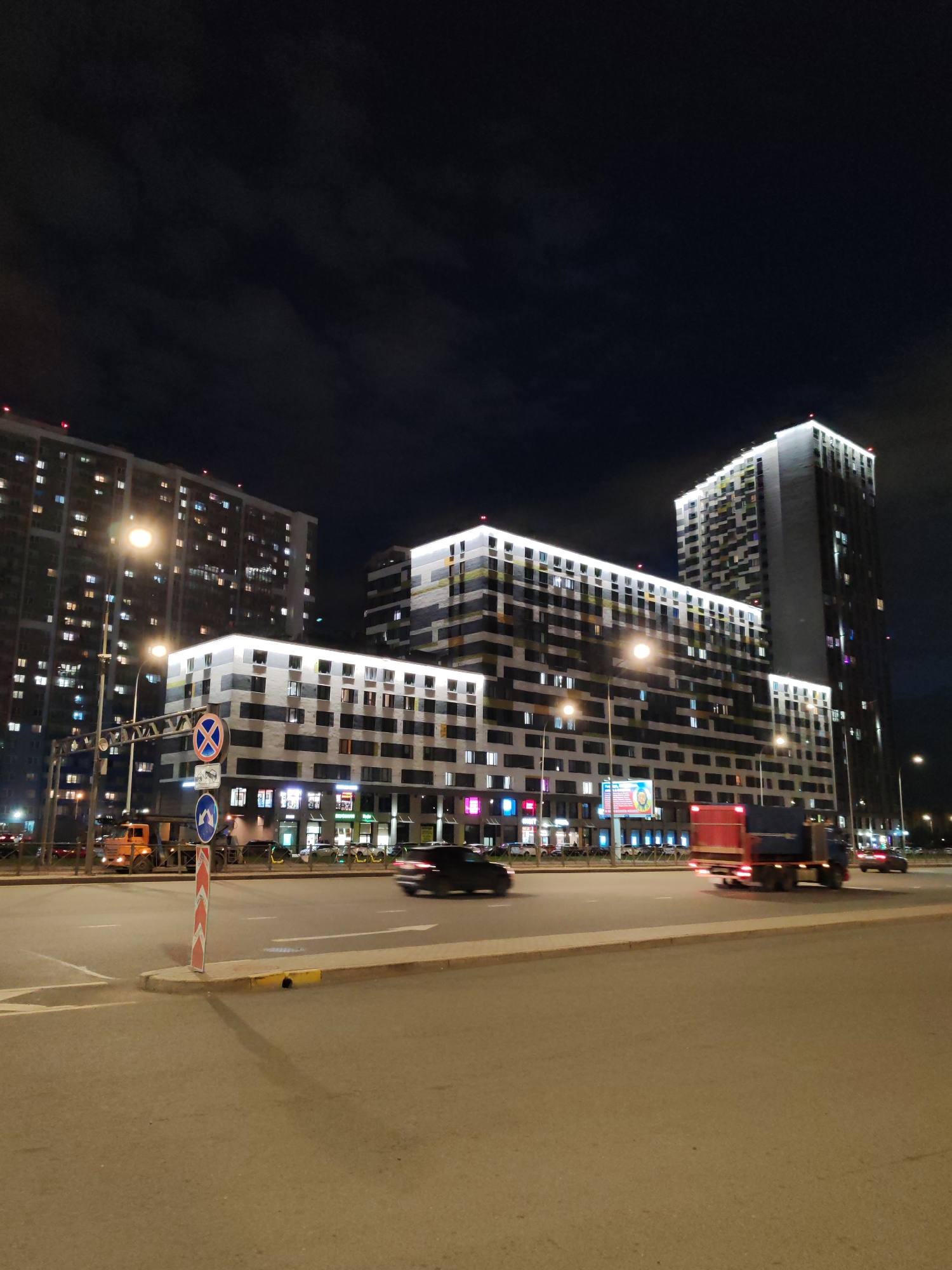 This nighttime image captures a bustling urban scene in what appears to be Saint Petersburg, Russia. The primary subjects are several large, modern apartment buildings alongside a multi-lane road, illuminated by a myriad of artificial lights under a dark sky.
The background is dominated by three prominent high-rise residential buildings. The structure on the far left has a darker facade with numerous glowing windows, indicating active units. The central and right buildings feature distinct contemporary designs with patterned facades composed of grey, black, yellow, and white panels. These buildings are dramatically lit by white LED strip lighting tracing their rooflines and some intermediate levels, enhancing their modern aesthetic. Many windows across all buildings are lit, suggesting residential occupation. At the base of the central building, what appear to be storefronts or commercial spaces are visible, along with a large, illuminated billboard-like sign containing indistinct text.
In the foreground and midground, a wide asphalt road stretches across the frame. Vehicles are present and in motion; a dark car on the left and a red and blue truck on the right show motion blur, indicating ongoing traffic. A yellow/orange utility or construction truck is also visible near the left building. The road features white lane markings, including a clear right-pointing arrow in the foreground. A prominent lamppost with two bright lights casts illumination over the street. In the foreground, a red and white striped barrier pole supports two circular blue Russian road signs: one indicating "No Stopping" (a red cross on a blue background) and another showing a mandatory left turn arrow. A concrete median with yellow painted curbs separates parts of the road.
The scene conveys a sense of a lively, modern city after dusk, with well-lit infrastructure and active urban life.