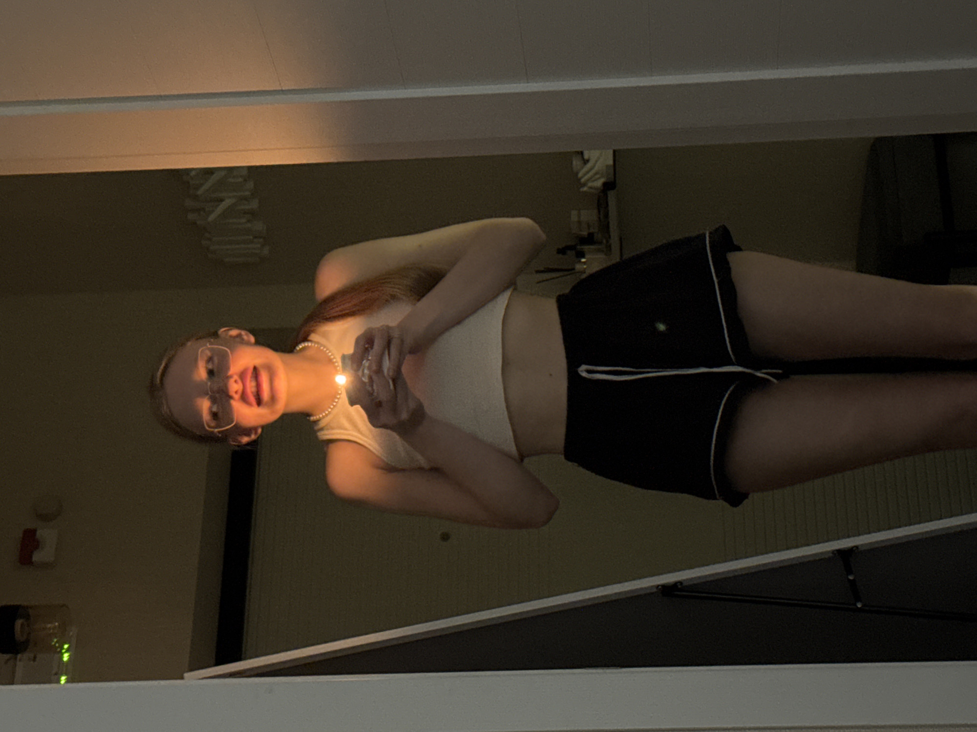 An image, likely taken in Perm, Russia, depicts a young person indoors, rotated 90 degrees clockwise from a natural upright view. The subject, who appears to be female, has light brown or reddish hair and wears glasses, a light-colored crop top or tank top, and dark shorts with white trim. A pearl necklace adorns her neck. She is holding a small, bright light source, possibly a candle or a tiny lamp, close to her face, illuminating her features and reflecting off her glasses. Her mouth is slightly open, and she has a playful or happy expression.
The background indicates an indoor setting, likely a room or hallway. The ambient lighting is dim, suggesting it is nighttime or a low-light environment. On the left wall, a red device resembling a fire alarm or emergency switch is visible. Below it, a clear container holds glowing green elements, possibly decorative lights or a functional electronic device. In the upper part of the image, a modern light fixture or chandelier with multiple rectangular elements is suspended from the ceiling. A white horizontal structure crosses the frame, possibly a window ledge or architectural feature. No discernible text is visible in the image.