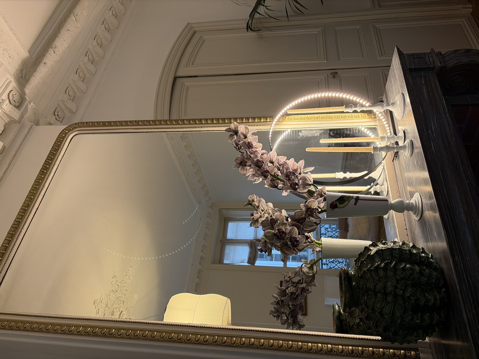 This image depicts a static interior scene within what appears to be a classic Haussmannian apartment in Paris, France. The primary focal point is a large, rectangular mirror with an ornate, gilded frame, positioned vertically against a light-colored wall. The mirror reflects a high ceiling adorned with intricate white plaster molding and a light-colored, possibly fabric, pendant lamp. A window with visible exterior ironwork, likely a balcony railing, is also reflected in the mirror.
Below the mirror, a dark, potentially marble, fireplace mantelpiece serves as a display surface. On the left side of the mantelpiece rests a dark green, textured vase with a rugged, possibly organic, surface pattern. To its right, two tall, slender white vases hold multiple stems of pale purple orchids. One of these vases features a small, colorful bird illustration. Further to the right, three white candlesticks of varying heights stand on the mantelpiece, each holding an unlit, cream-colored candle. Behind these candlesticks, a circular LED ring light, emitting a bright white glow, is visible.
To the right of the main mirror, a set of white double doors with an arched transom are visible, consistent with typical Parisian architectural features. A hint of a green, leafy plant is partially visible in the far right of the frame. The overall lighting suggests an indoor setting with both artificial and natural light sources contributing to the scene's illumination.