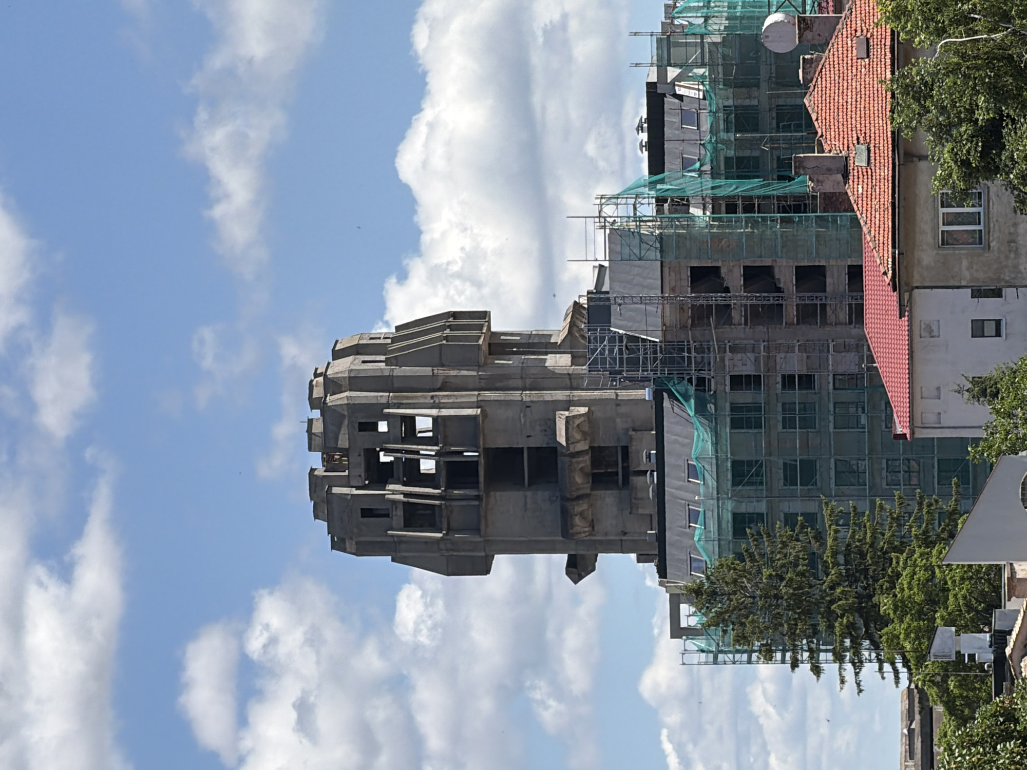 In Shumen, Bulgaria, a multi-story building, appearing to be under construction or renovation, is depicted against a bright blue sky with scattered white clouds. The building's upper section features a distinctive, fortress-like concrete structure with numerous openings. The middle sections are covered in green netting and scaffolding, suggesting ongoing work. A tall evergreen tree is visible on the left side, adjacent to the building, with dense foliage. In the foreground, several smaller, older residential buildings with red-tiled roofs are visible, some partially obscured by trees. The overall scene is an urban environment under a clear sky, highlighting a significant construction project.