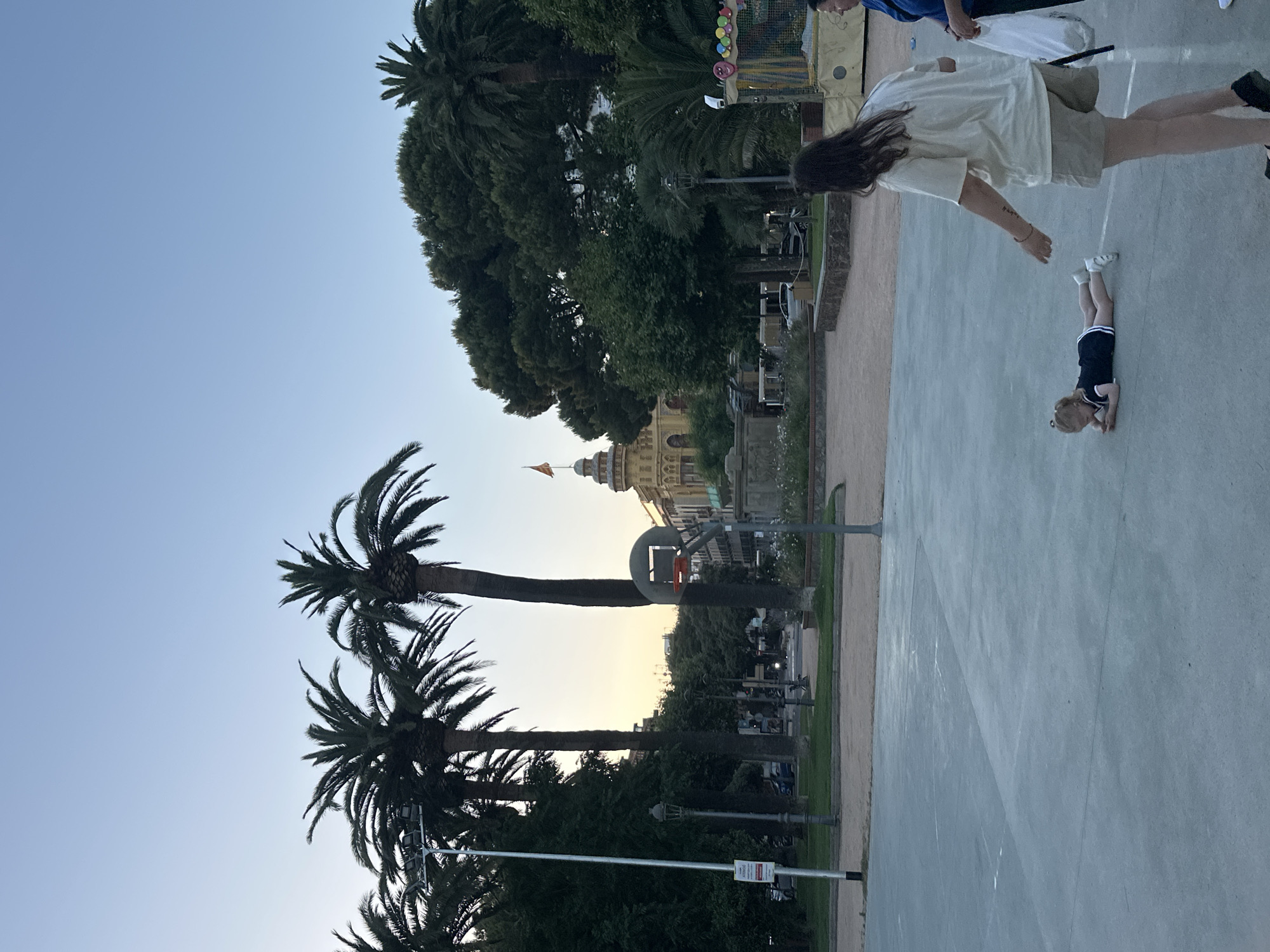 This is an outdoor shot of a park scene in Sant Feliu de Guíxols, Spain, during daylight hours, likely late afternoon or early evening given the lighting. In the foreground, a child wearing a dark blue dress with white trim is lying face down on a grey basketball court. To the right of the child, a woman with long dark hair and wearing a light-colored t-shirt and beige shorts is standing, facing the child with her right arm extended. Another woman, partially visible on the far right, is also present.
The background features several tall palm trees and other dense green foliage, typical of a Mediterranean climate. Behind the trees, a distinctive yellow building with a domed roof and a flag flying from its pinnacle is visible. A basketball hoop with a red rim is positioned on the court. The park appears to have walking paths, benches, and streetlights. In the far right background, a colorful, inflatable structure, possibly a bouncy castle, is present. The sky is a clear light blue.