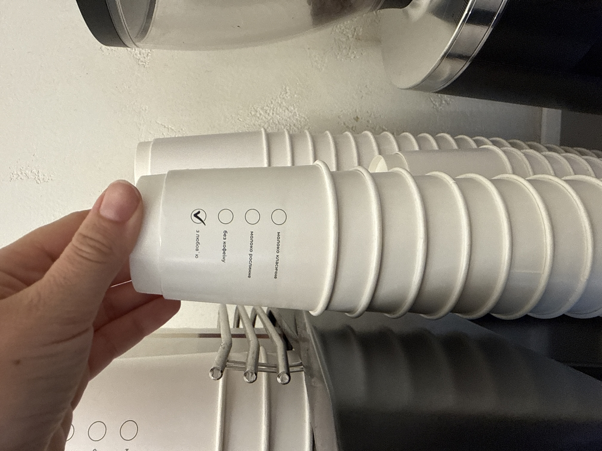 A high-angle, close-up shot captures a person's hand interacting with a self-service coffee station in Uzhhorod, Ukraine. The hand, with manicured nails, is positioned above a stack of white paper cups, either selecting or placing the topmost cup. The cups feature black Ukrainian text and circular checkboxes arranged vertically. Visible text, read from top to bottom on the cup being held, includes "З собою" (To go) with a checkmark, followed by unchecked options: "Без кофеїну" (Decaf), "Молоко рослинне" (Plant milk), and "Молоко класичне" (Classic milk). To the immediate right, a taller stack of identical white paper cups is visible. Further to the right, a black and silver coffee grinder or dispensing machine with a transparent hopper containing dark coffee beans is present. On the far left, a portion of a white dispenser or machine with circular buttons is visible, adjacent to a metal rack holding more white cups. The background consists of a light-colored wall with textured, possibly flaking, paint.