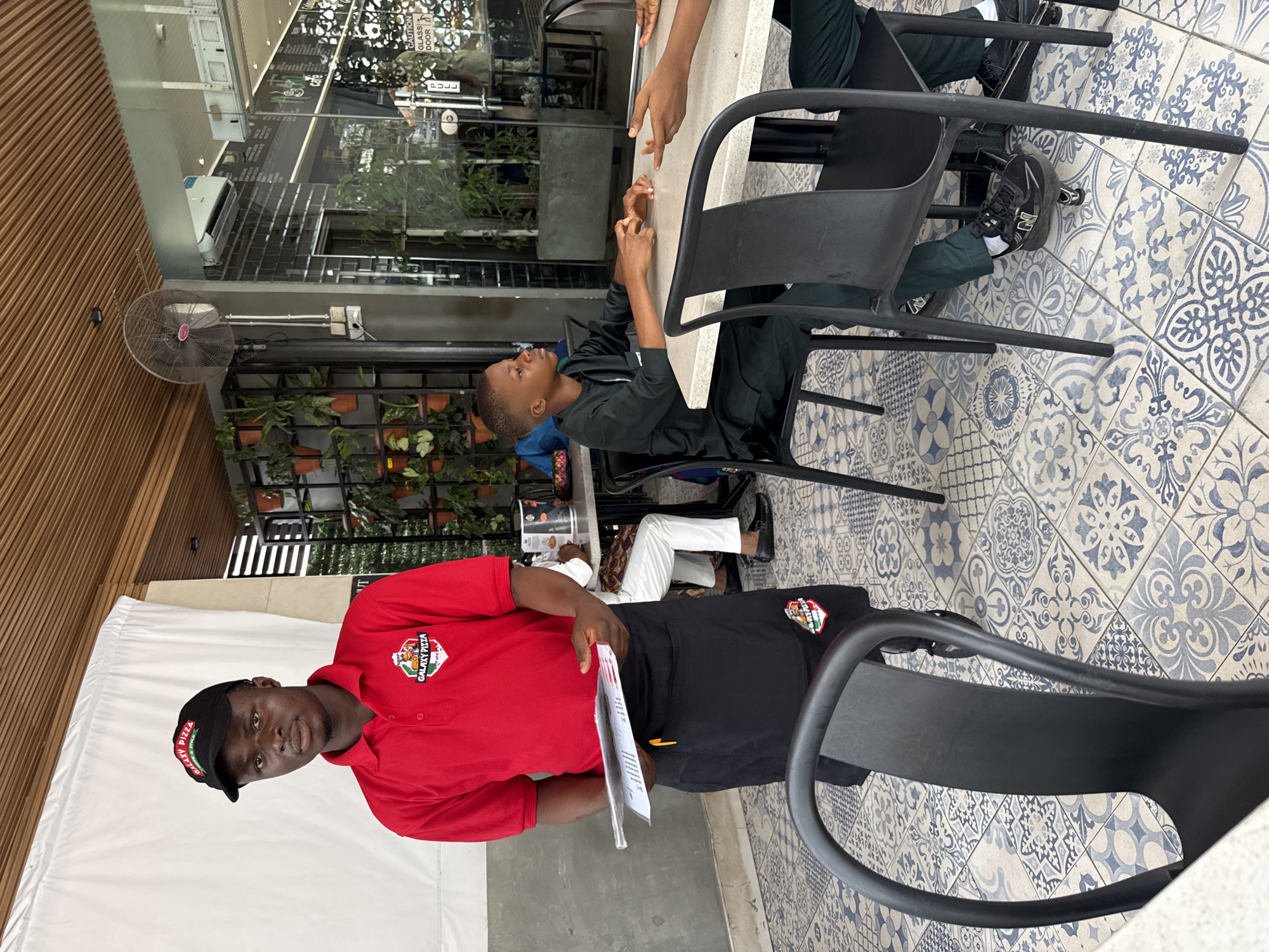 The image shows an outdoor restaurant setting in Abuja, Nigeria. The floor is tiled with blue and white patterned tiles. A Galaxy Pizza employee in a red shirt, black apron, and Galaxy Pizza cap stands holding a menu. Two young customers sit at a table, one dressed in dark green. There are black chairs around the table. Planter boxes with greenery are visible in the background along a glass wall. A fan is mounted on the ceiling.