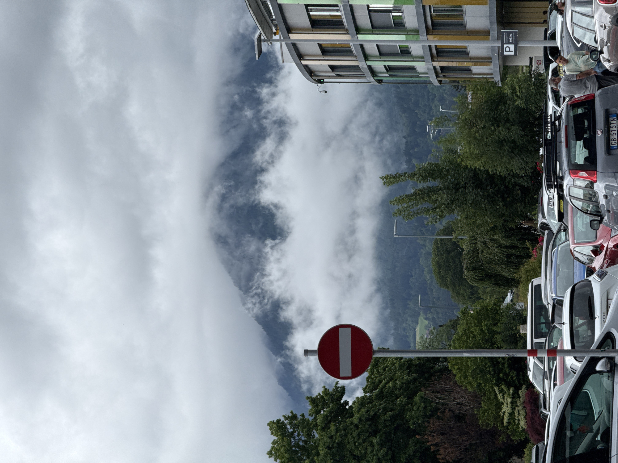 An overcast day with low-lying clouds obscures the upper portions of a mountain range behind a multi-story building with a curved facade. In the foreground, a "no entry" traffic sign is prominent, with a busy parking lot filled with various cars. Two men are visible near the cars, one leaning on a vehicle and the other standing beside it, engaged in conversation. The architecture suggests a modern urban setting, possibly a commercial area or office complex. The overall atmosphere is subdued due to the weather conditions.