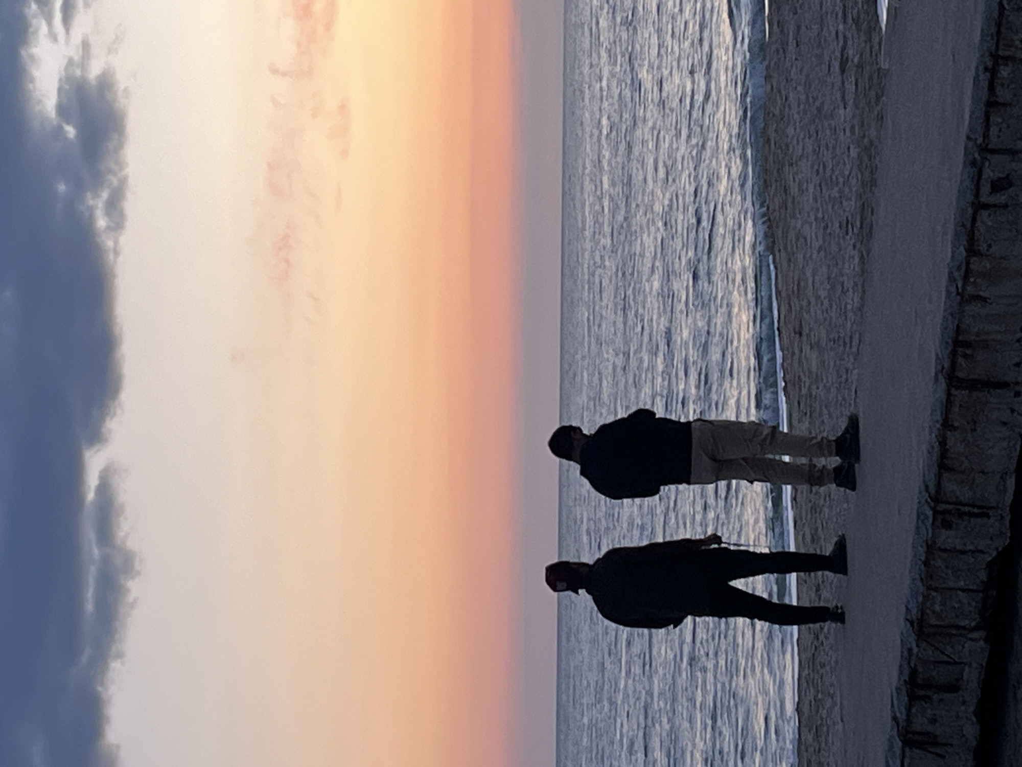 Two people stand on a stone structure next to the sea at sunset. One person wears a dark jacket and pants, and the other wears a dark jacket and light pants. The sunset is visible in the sky over the sea, with colors ranging from blue to orange. The stone structure appears weathered, and the sea is slightly choppy. This is the city of Alanya, Turkey.