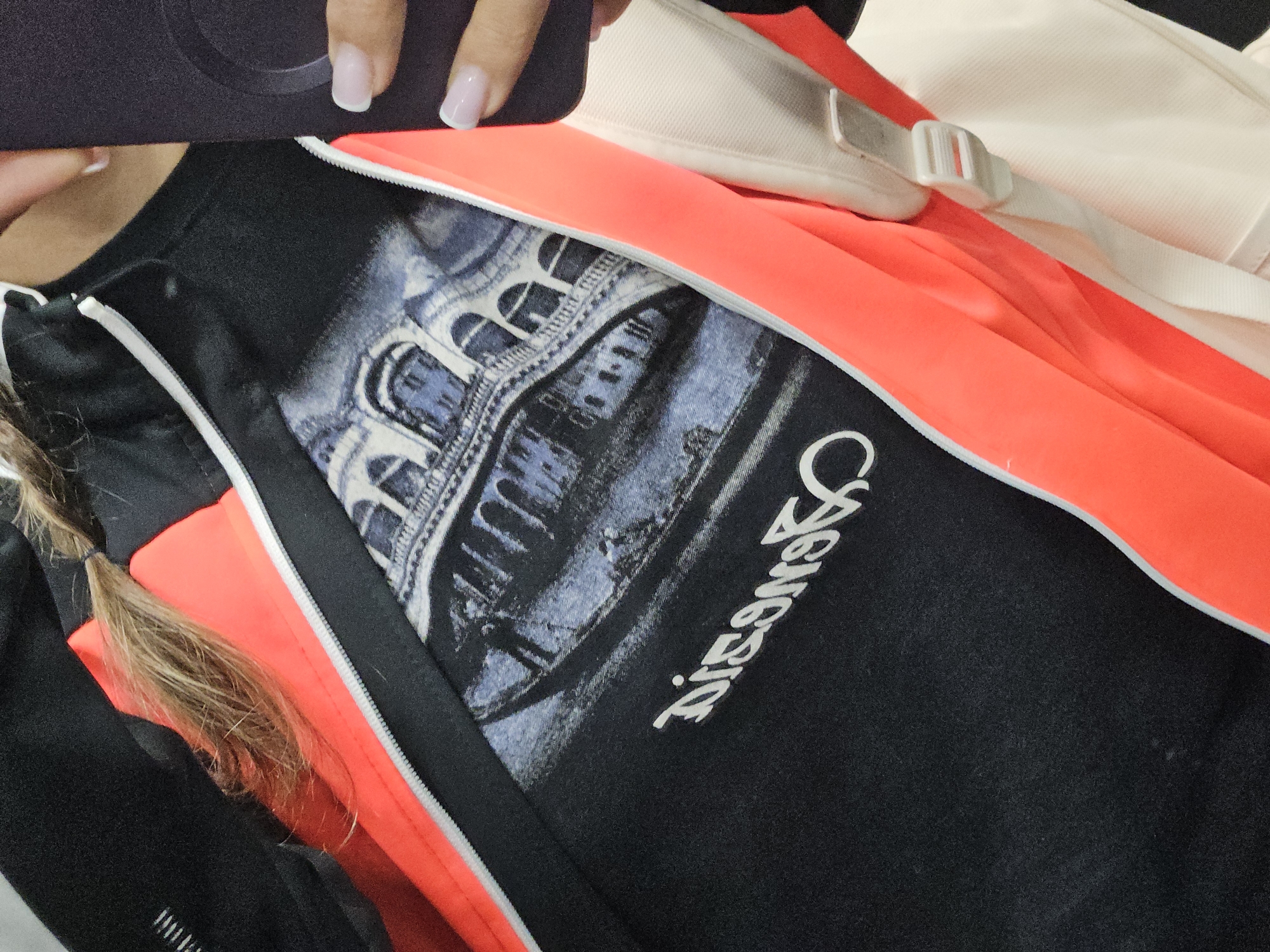 A close-up shot presents a person holding a phone up to capture a selfie. The subject's face is obscured, with only their manicured hand and the phone visible at the top of the frame. They are wearing a black t-shirt imprinted with an image of an arched building and the word "GENESIS" printed vertically in white font. The t-shirt is partially covered by an open athletic jacket, which is black with a white zipper line and bright orange lining. A tan backpack strap rests diagonally across the top right of the frame. A ponytail is partially visible at the left side of the frame.