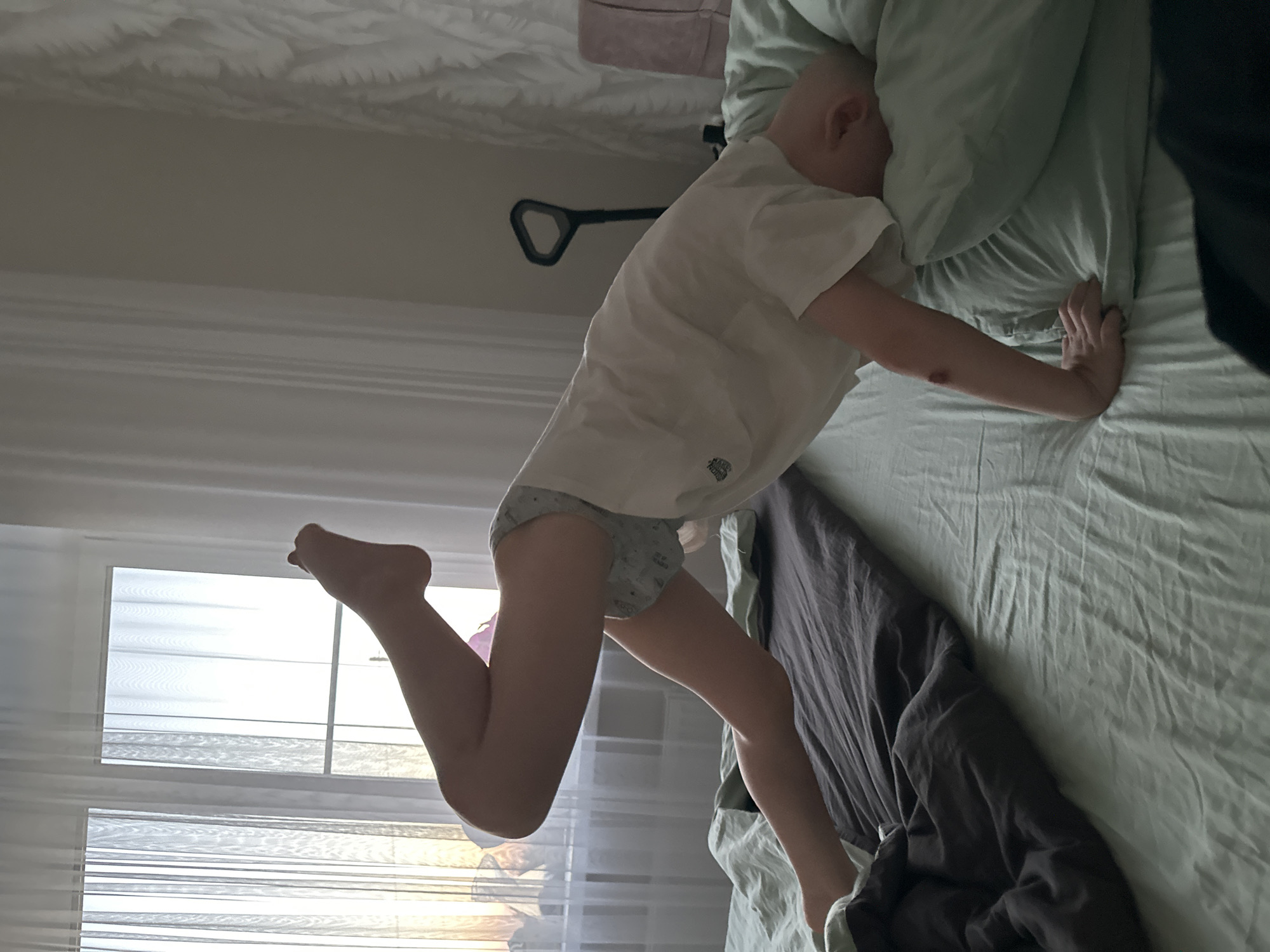 A person, possibly a child, is in bed. The person is wearing a white t-shirt and patterned underwear. They are lying on their stomach with their head on a pillow and one arm extended to the side. One leg is bent at the knee and raised in the air. The bed is covered in light green sheets and a dark gray blanket. A window with blinds is visible in the background. The location is possibly Прикамский, Russia.