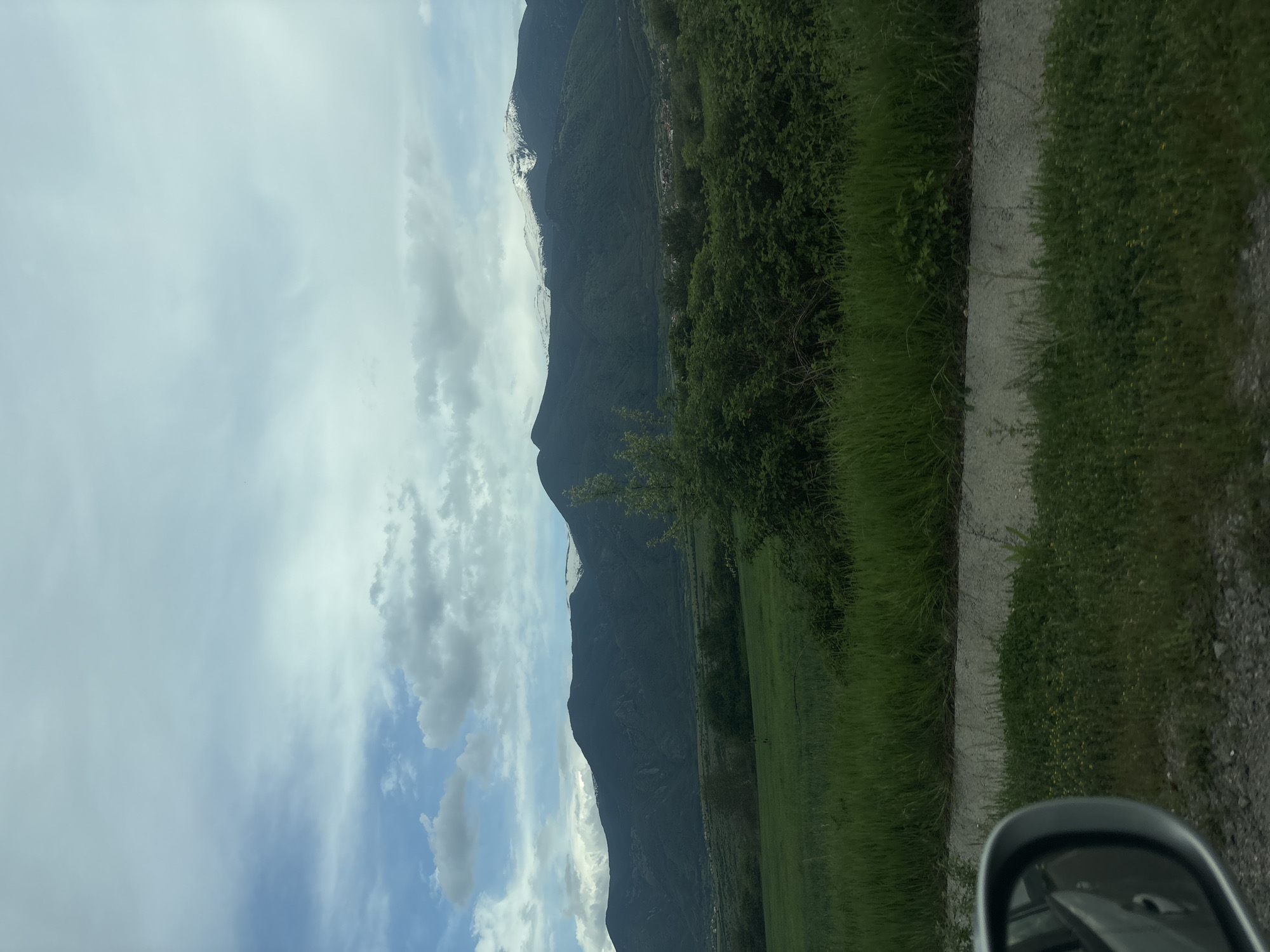 The image shows a landscape with a mountain range under a cloudy sky. Snow is visible on the peaks. In the foreground, there is a grassy field and a dirt road. A side view mirror is visible in the lower right corner. There is no indication that the image is in Yahinovo, Bulgaria. There are no people present in the scene.