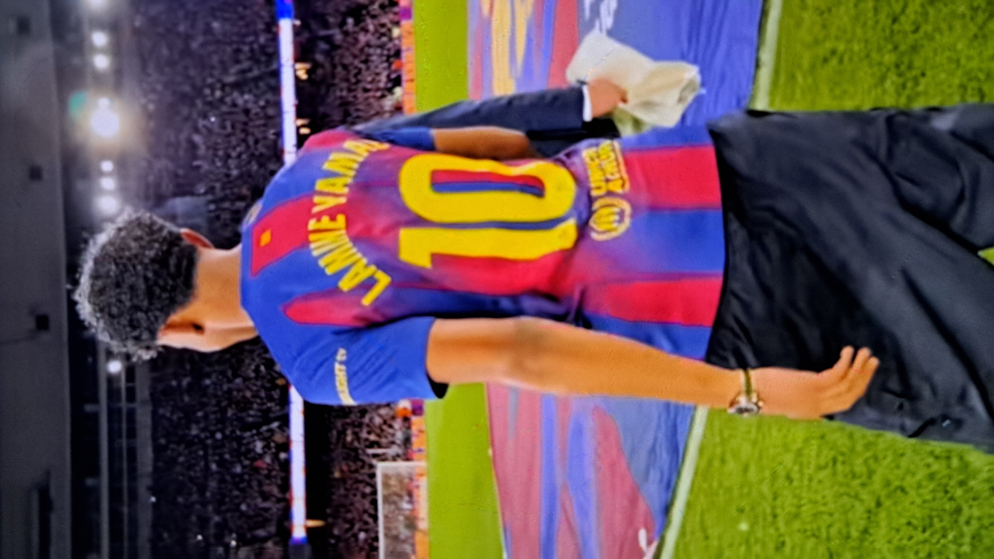 A young male, seen from the back, stands on a green grassy field, likely a football pitch. He wears a blue and red striped jersey with "LAMINE YAMA" and the number "10" in yellow. The jersey also has a small yellow UNICEF logo and a "BJK TV" logo on the left shoulder. He has dark, curly hair and is wearing black cargo pants and a watch on his left wrist. He holds a white cloth or towel in his right hand, draped over a dark jacket. The background shows a large crowd in stadium seating, illuminated by bright lights. The field has a large blue and red tarp. The image is framed such that the person is the primary subject, with the stadium and field serving as the setting. There is no information available to confirm the location as Payagaboh, Indonesia.