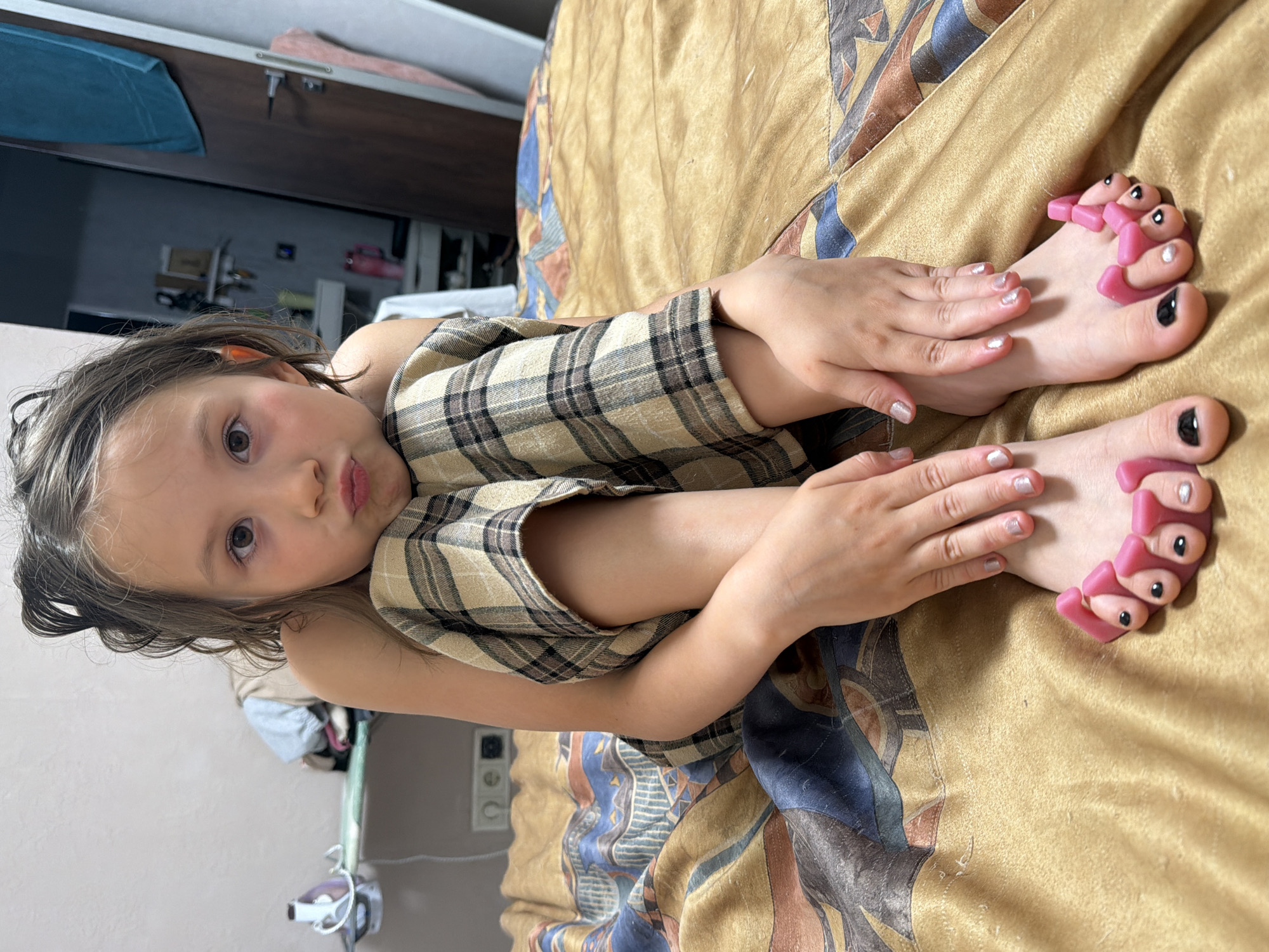 A young girl with light brown hair pulled into messy buns sits cross-legged on a bed covered with a textured golden blanket with a patterned accent. She is wearing a plaid top and pants. Her fingernails are painted with a light shimmery polish, and her toenails are painted with black and silver polish. Pink toe separators are placed between her toes. The background appears to be an indoor setting, possibly a bedroom, with a wall, an ironing board with an iron, and a doorway visible. The scene suggests a casual, domestic environment. The location is the city of Perm, Russia.