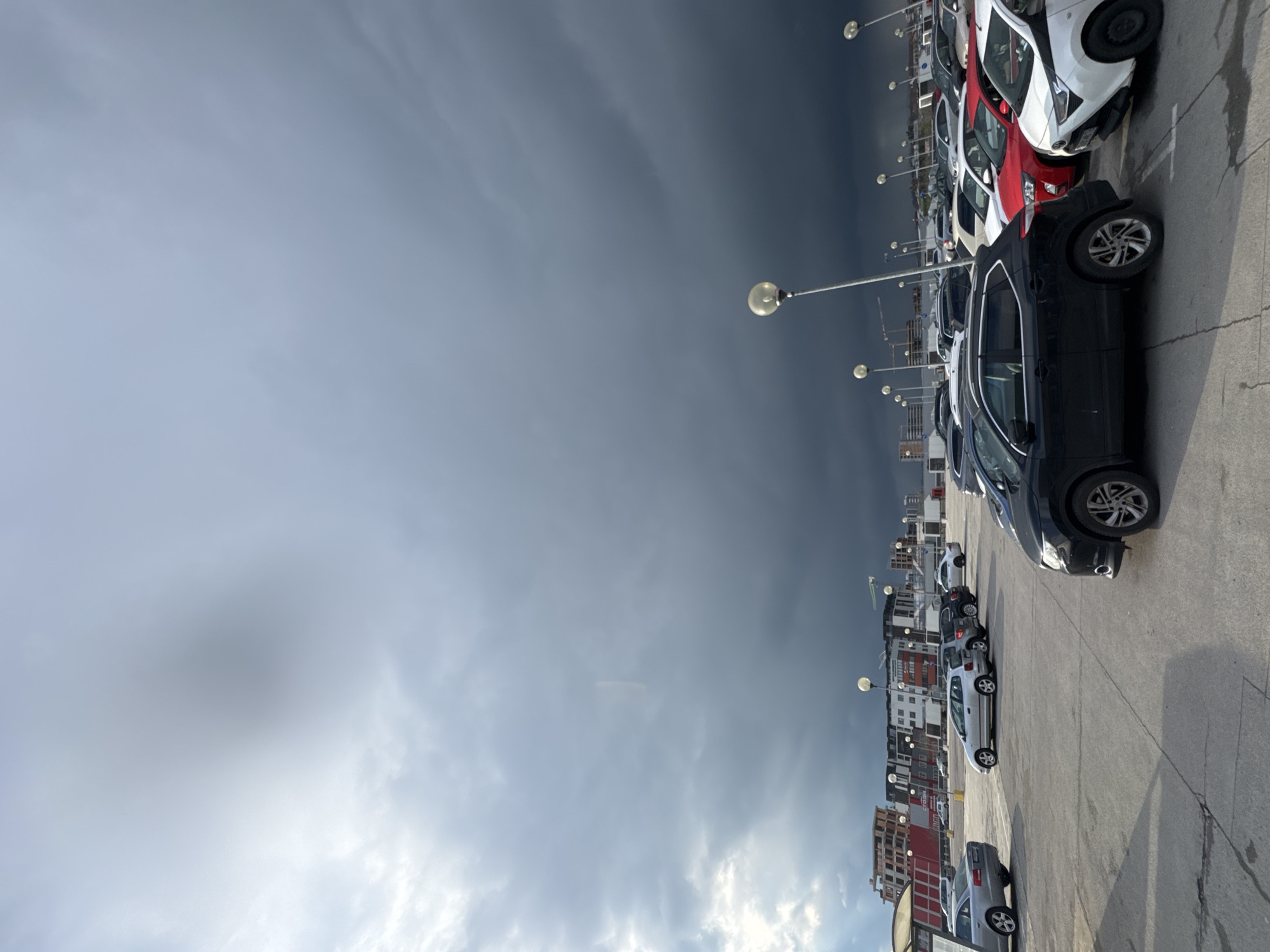 The image shows a parking lot in Burgas, Bulgaria, filled with rows of cars parked on asphalt. The cars are various colors including white, red, black, and silver. In the background, urban buildings can be seen. Overhead streetlights are visible along the parking rows. The sky is overcast and dark.