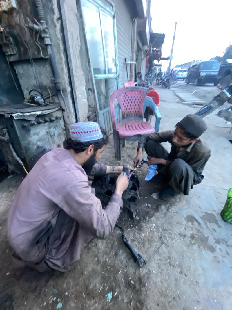 This image depicts two men working on a heavy metal component, likely an engine block, in what appears to be a street-side workshop in Kandahar, Afghanistan. The scene is set on a rough, unpaved or worn concrete surface scattered with dirt and debris, with an industrial-looking background.
The man on the left, distinguished by a beard and a light-colored skullcap with red and white patterns, wears a light brown or dusty purple long-sleeved tunic (shalwar kameez). He is crouched low, intently focused on the metal object, using what appears to be a hammer and chisel or similar tools. A long, dark tool lies on the ground beside him.
Crouching to his right is a younger individual, possibly a boy or young man, wearing a dark tunic and trousers with a dark skullcap. He is also actively engaged with the same metal component, with his hands busy, one of them possibly covered by a light blue cloth or glove. Both individuals are deeply concentrated on their task.
In the background, an old building or shop front with a large window and what seem to be industrial fixtures like pipes and machinery are visible on the left wall. Behind the men, a red and blue plastic chair stands partially visible. The further background shows a blurry urban streetscape with vehicles, including cars and motorcycles, and more buildings, suggesting a bustling yet utilitarian environment.
The lighting appears soft and diffused, indicating either early morning or late afternoon. The overall atmosphere is one of diligent manual labor in a practical, functional setting characteristic of roadside repair operations. No distinct text is visible in the image.