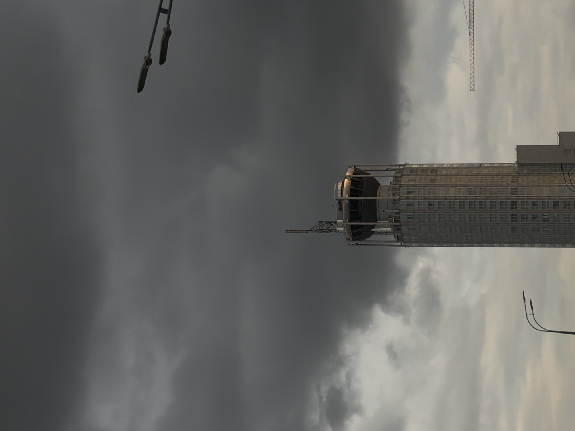 The image presents a tall, modern building set against a striking and dramatic sky. The building, likely a skyscraper, is slender and characterized by multiple floors with visible windows. Its upper portion features a distinctive, rounded structure, possibly an observation deck or architectural accent, with scaffolding or framework around it.
The sky is the dominant element, displaying a sharp contrast. The left side is dominated by dark, heavy grey clouds, suggesting an impending storm or very dense overcast conditions. Towards the right, the clouds lighten considerably, revealing more diffuse grey and white formations, indicating a break in the weather or the sun attempting to pierce through.
In the upper right area of the lighter sky, the arm of a construction crane is visible, hinting at ongoing development in the urban landscape. Two streetlights or similar utility fixtures are prominent in the upper left corner, appearing to hang downwards. Another streetlight structure is situated in the lower right, curving upwards.
No people are visible, and no specific activity is depicted. The lighting on the building suggests either early morning, late afternoon, or a heavily overcast day where some light is present. The overall scene conveys a somewhat moody and imposing atmosphere due to the dramatic sky. There is no visible text in the image.